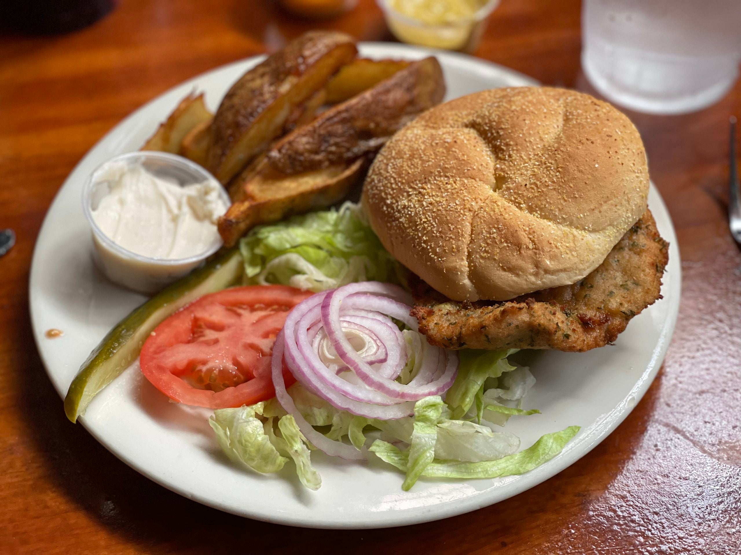 A plate showcasing one of the restaurant specialties: a chicken sandwich on a bun with lettuce, tomato, red onion, and pickle, accompanied by potato wedges and a cup of white dipping sauce, served on a wooden table.