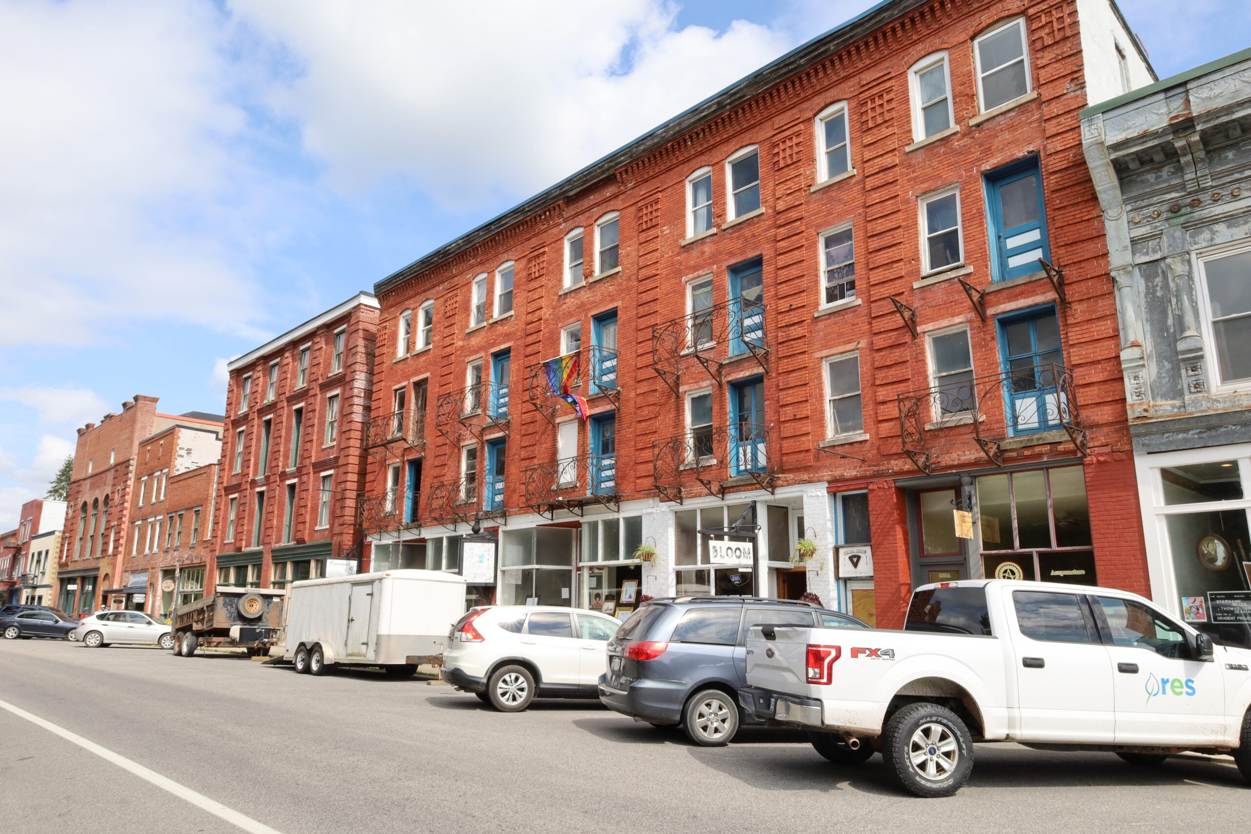 Brick buildings line a street with several parked cars and trucks. Storefronts and small businesses occupy the ground level, and blue sky with clouds is visible above.