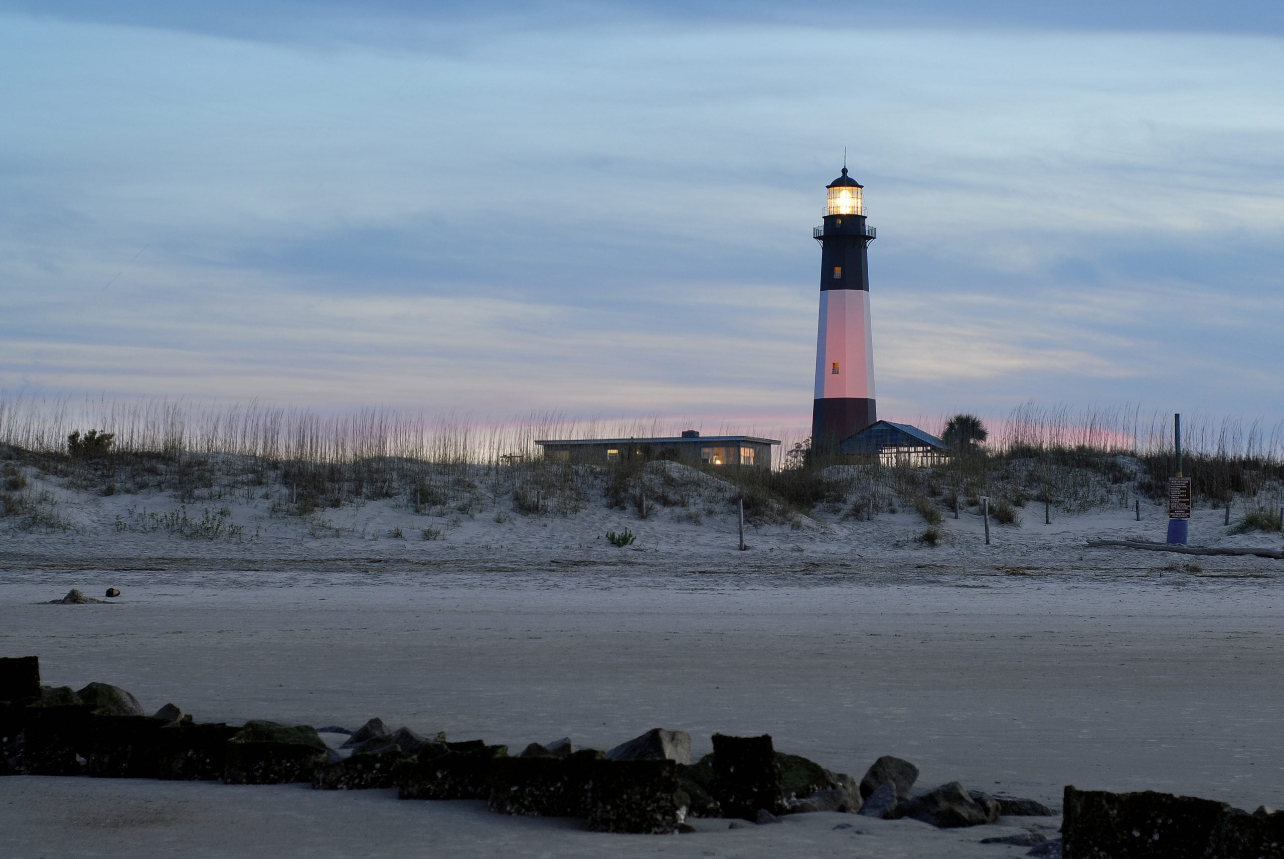 A lighthouse stands on sandy dunes at dusk, its light glowing against a blue and purple sky. Beach grass and a small building are nearby; rocks are visible in the foreground.