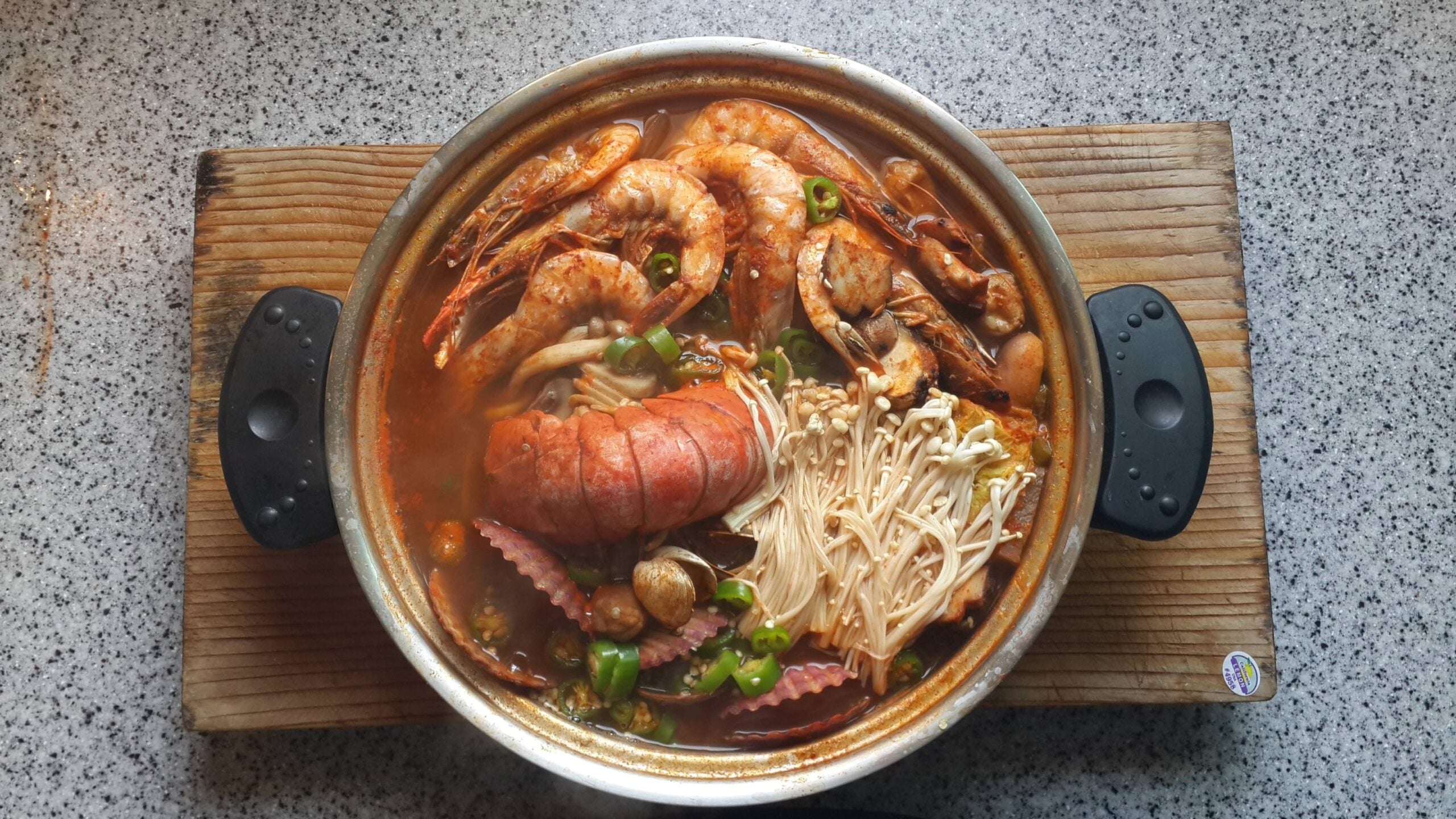 A pot of seafood soup with shrimp, lobster tail, octopus, mushrooms, and vegetables sits on a wooden board on a speckled countertop.
