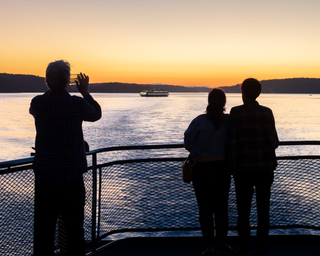 Three people on a ferry deck watch a sunset over calm water—one takes a photo while two stand close together. A distant ferry hints at trips where getting there is part of the adventure.