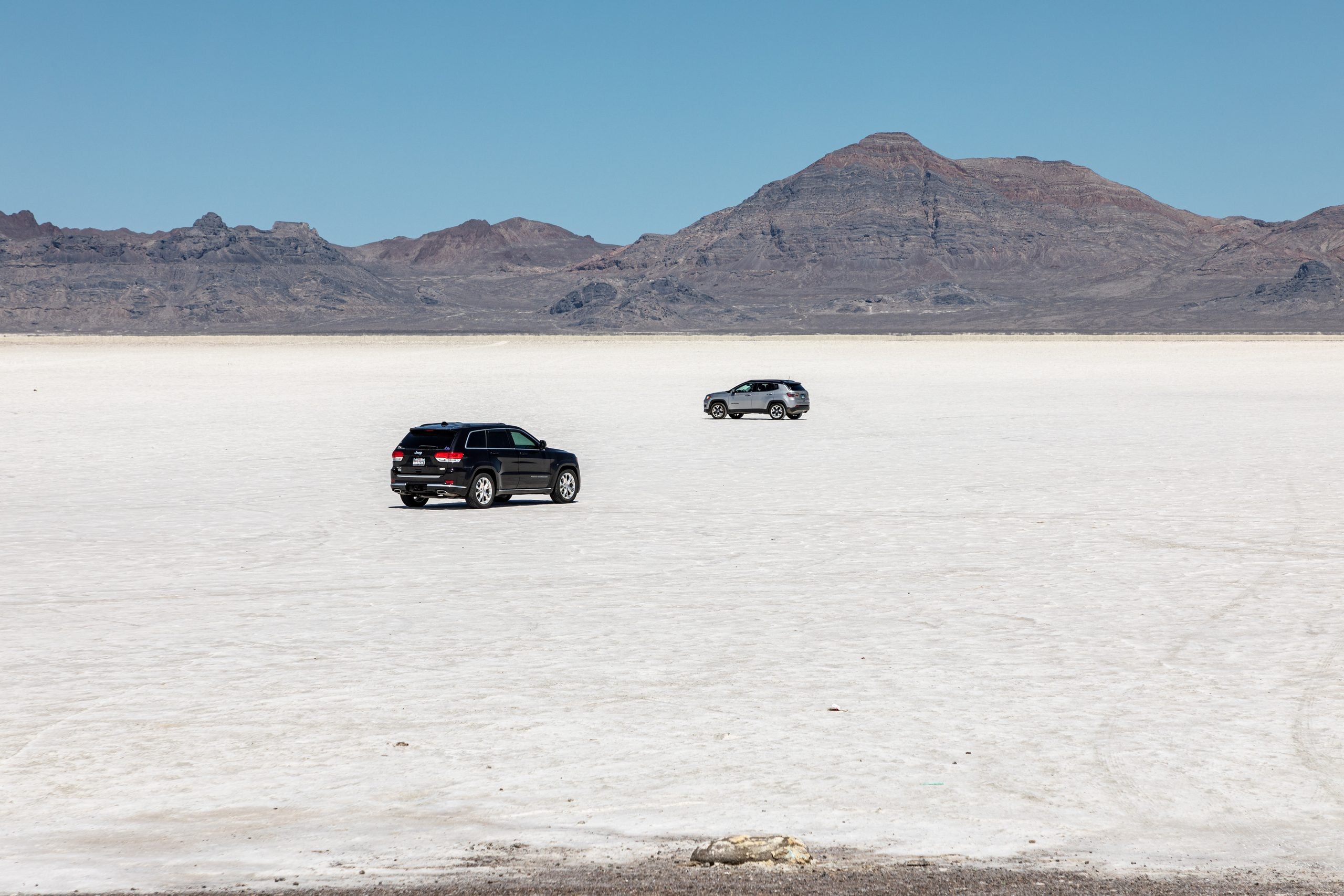Two SUVs drive across a vast, flat, white salt flat with rocky mountains in the background under a clear blue sky.