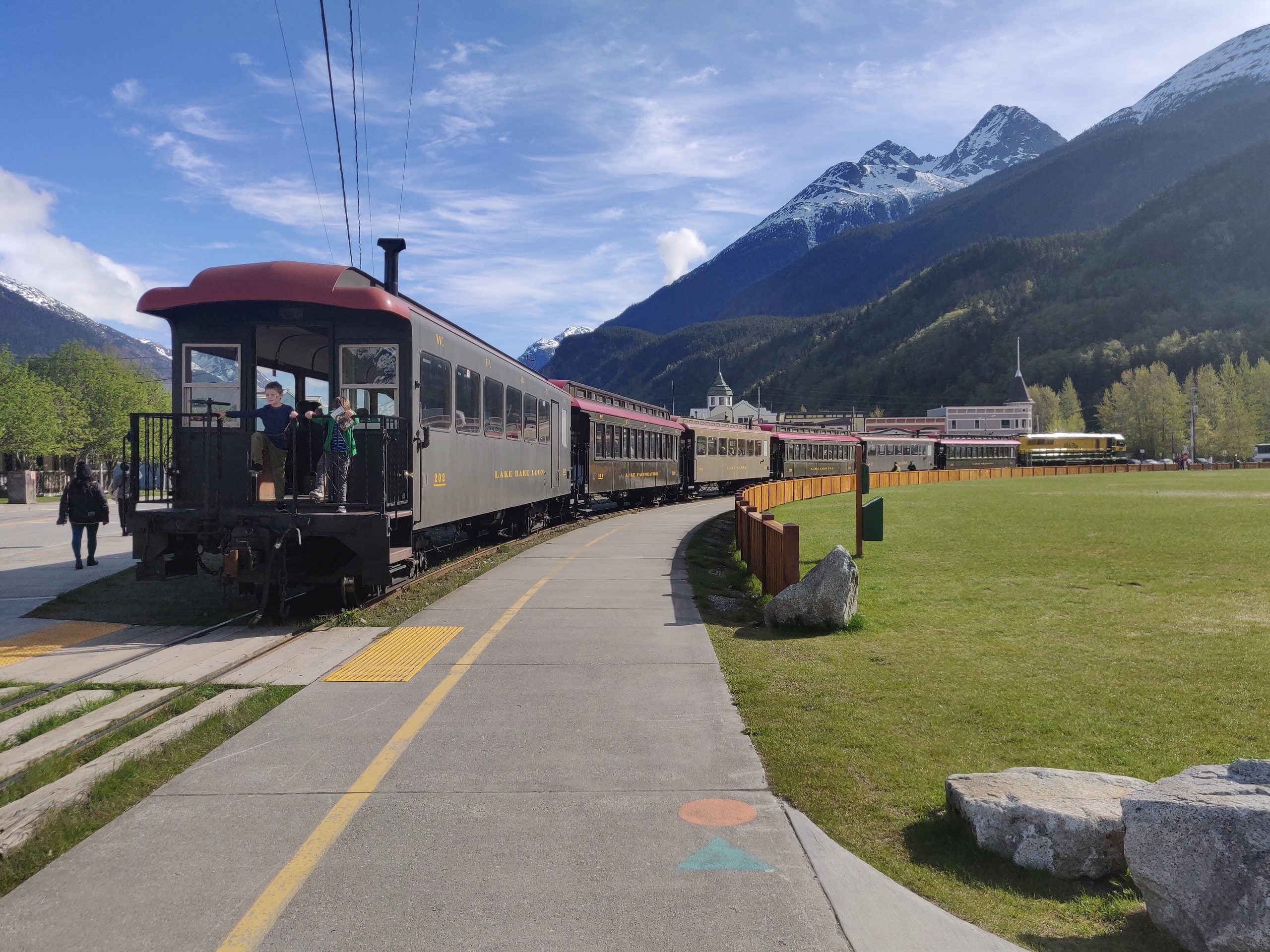 A vintage train with passengers boards at a platform beside a wide sidewalk, capturing the magic of trips where getting there is part of the adventure—mountains and buildings rise under a blue sky.