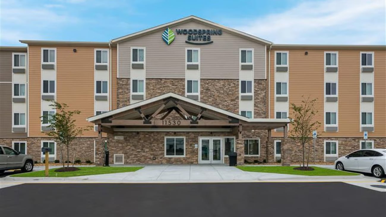 Four-story WoodSpring Suites hotel, part of well-known hotel chains, with a tan and stone exterior, many windows, and a covered entrance. Two small trees flank the walkway, and two cars are parked at the sides. Blue sky with some clouds overhead.