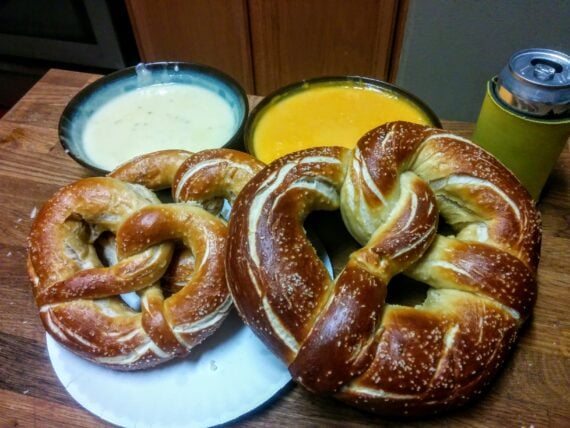 Two large, salted soft pretzels on a plate with bowls of cheese dips and a canned drink in a yellow holder on a wooden table.