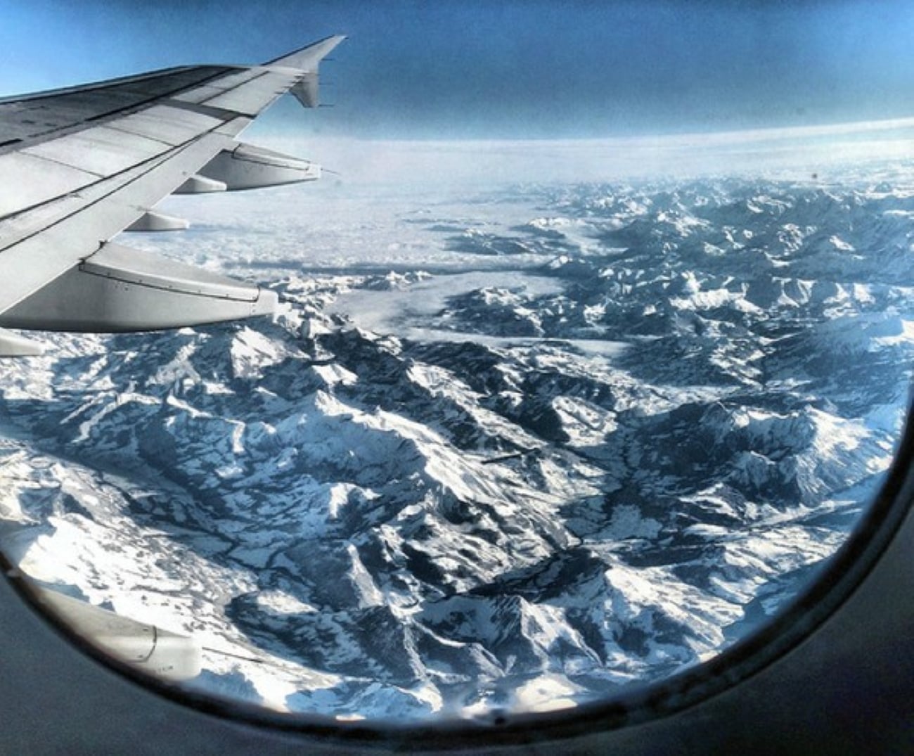 View from airplane windows showing the wing and snow-covered mountain ranges beneath a clear blue sky.