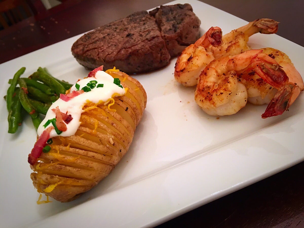 Grilled shrimp, a filet steak, green beans, and a baked potato topped with sour cream, cheese, bacon, and chives served on a white plate.