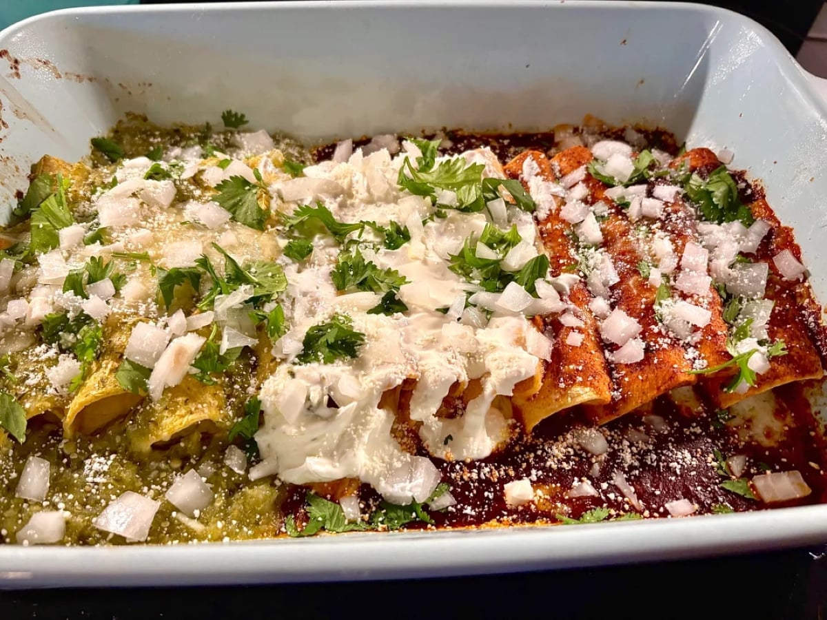A tray of enchiladas with green and red sauce, topped with chopped onions, cilantro, crema, and grated cheese.