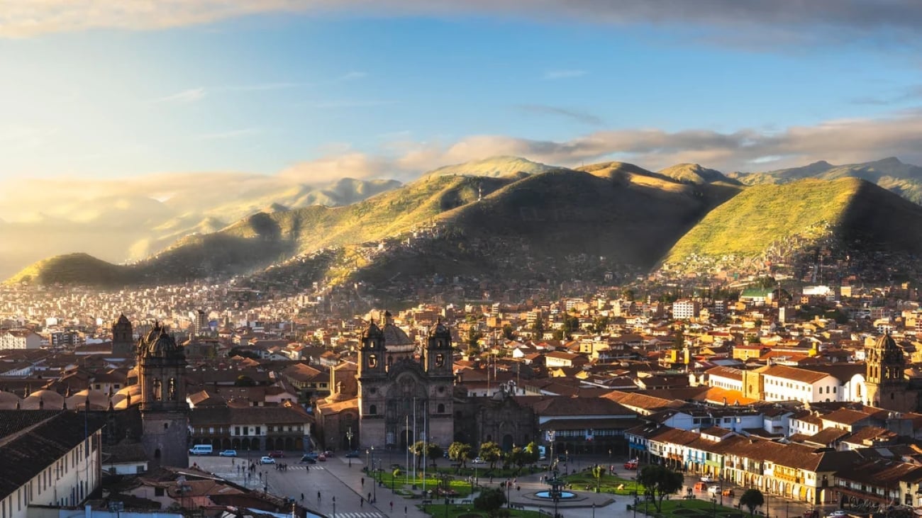 A panoramic view of Cusco, Peru, with historic buildings and churches in the foreground and sunlit green mountains in the background under a partly cloudy sky—a striking scene that contrasts with the bustling skylines of some of the Biggest Cities In The World.