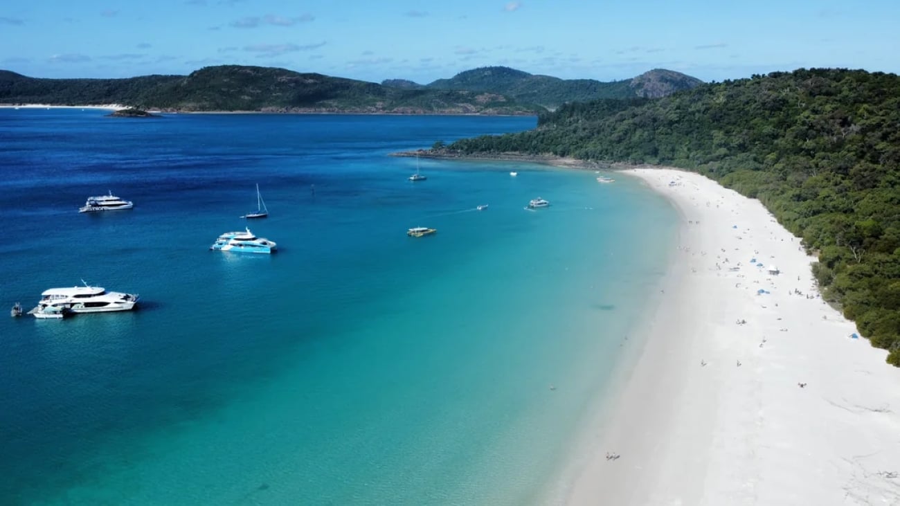 Aerial view of a tropical beach with white sand, turquoise water, anchored boats, and green hills in the background under a partly cloudy sky—an idyllic scene that captures the essence of sought-after tropical locations.