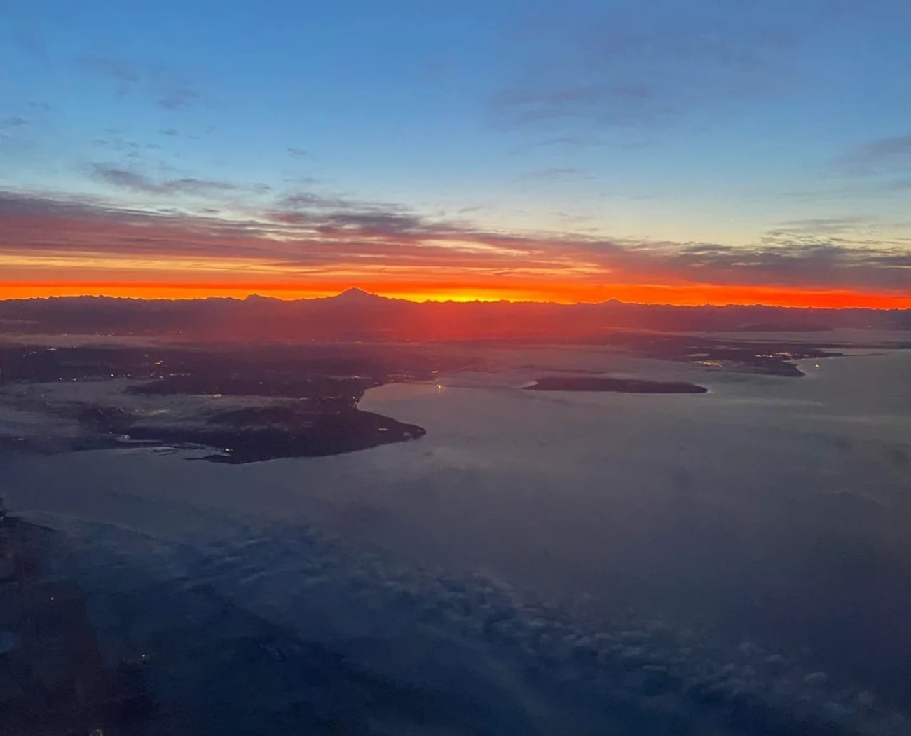 Aerial view of a coastline at sunrise or sunset, with dark blue water, scattered clouds, and a vibrant orange and red horizon beneath a mostly clear sky, as seen through airplane windows.