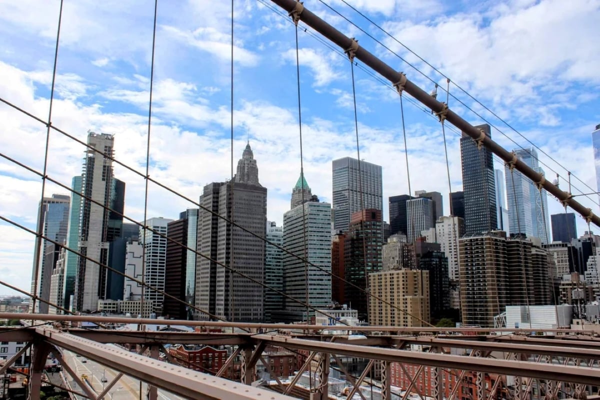 The New York City skyline viewed through the cables of the Brooklyn Bridge on a partly cloudy day. Tall skyscrapers rise against the blue sky.
