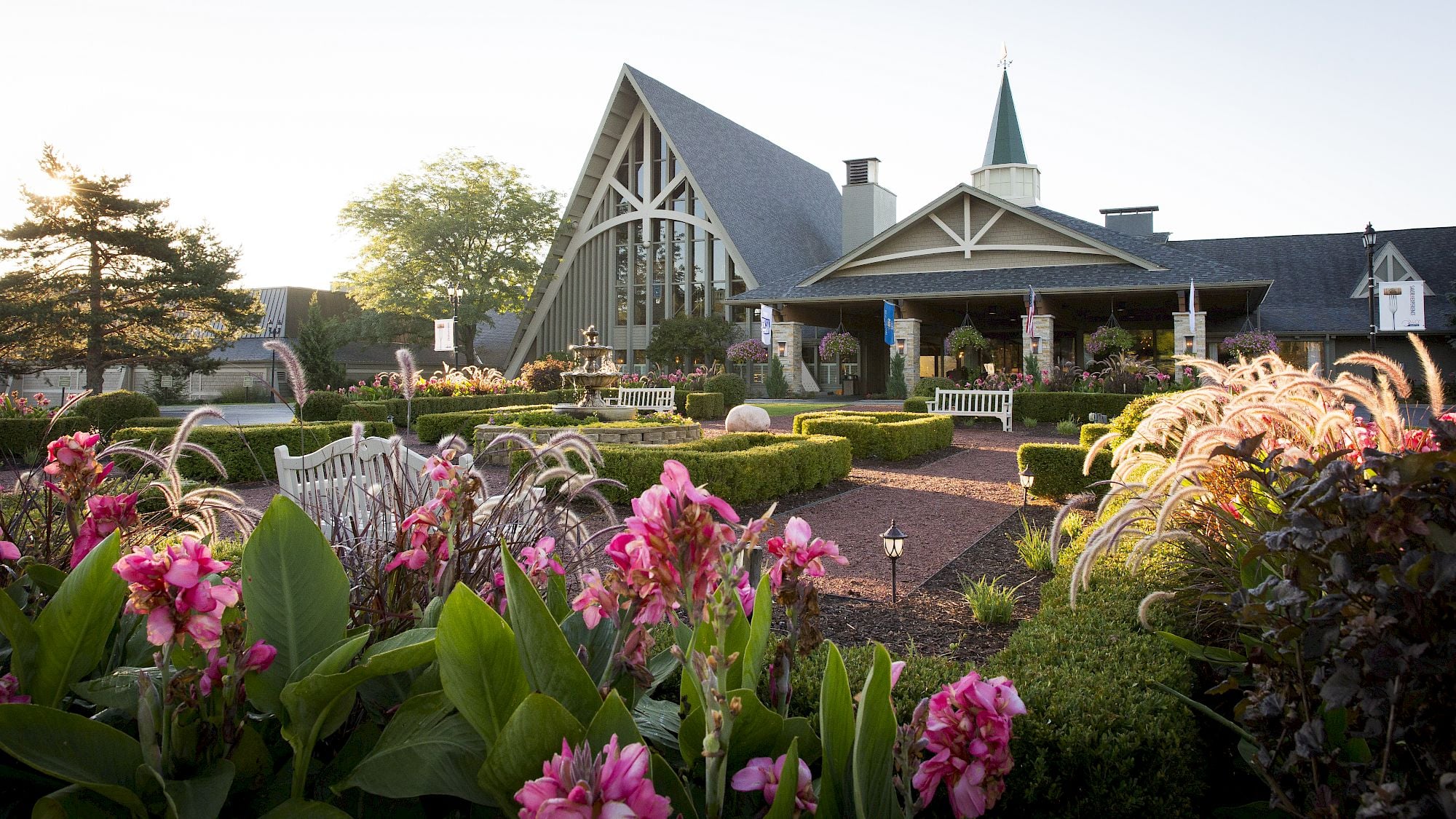 Flower garden with pink blooms in front of a large building with an arched window, green steeple, and benches—reminiscent of the tranquil grounds often found at peaceful resorts—all lit by soft evening sunlight.