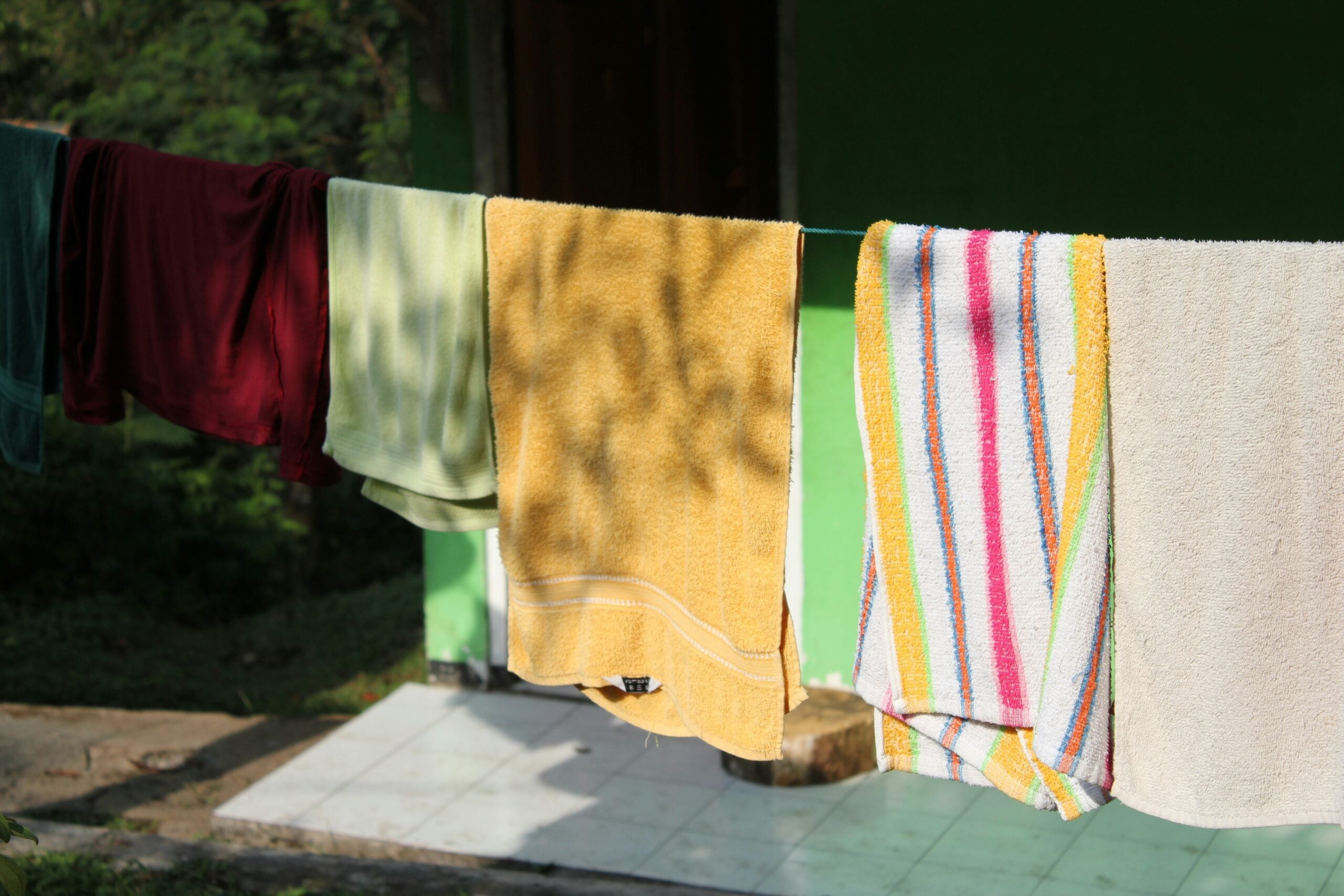 Several towels in different colors, including yellow, striped, and burgundy, hang on a clothesline outside in the sunlight near a green building.