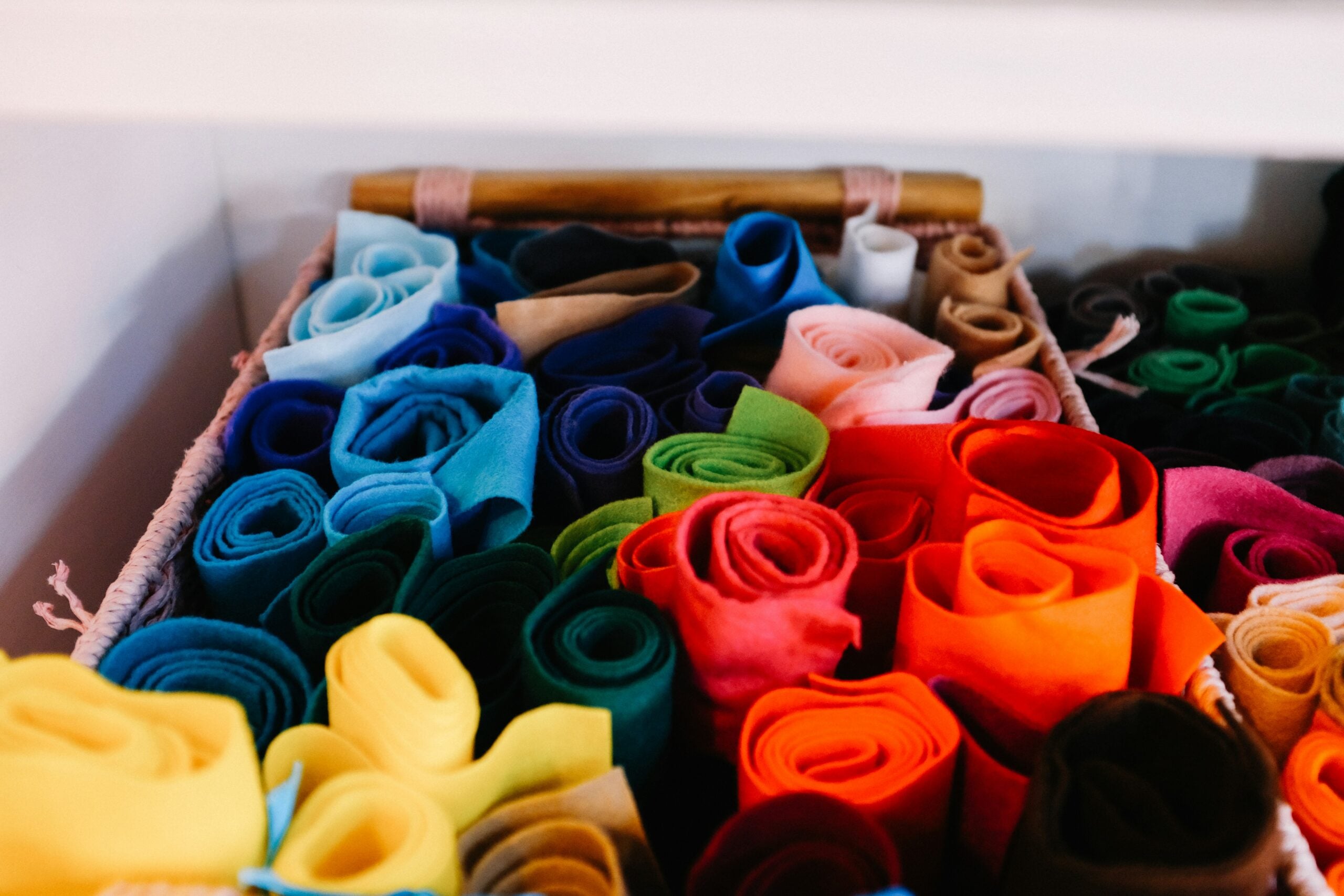 A basket filled with neatly rolled pieces of colorful fabric, organized by shades of blue, green, yellow, orange, red, and brown, on a shelf.