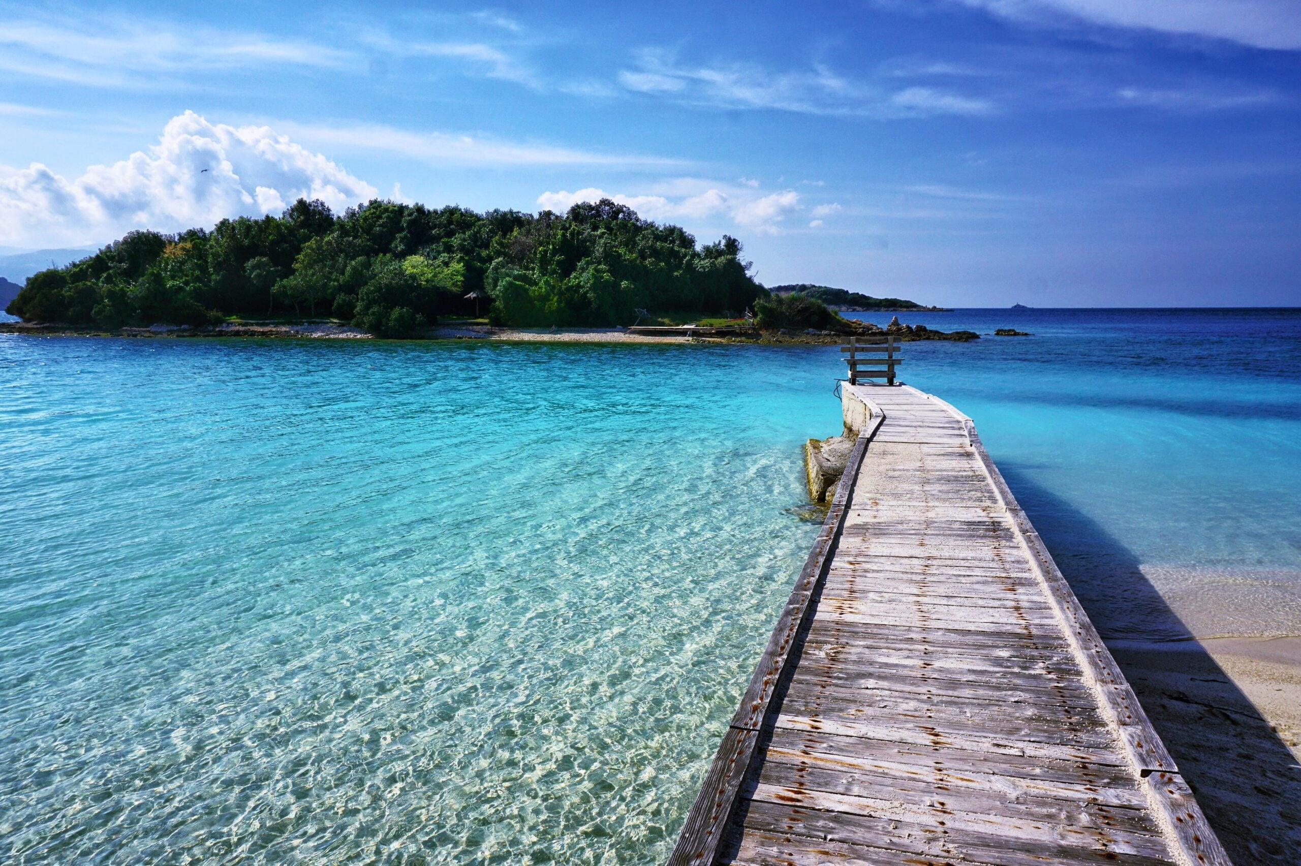 A wooden pier extends over clear turquoise water toward a lush, green island known for its incredible beaches, all beneath a bright blue sky with scattered clouds.