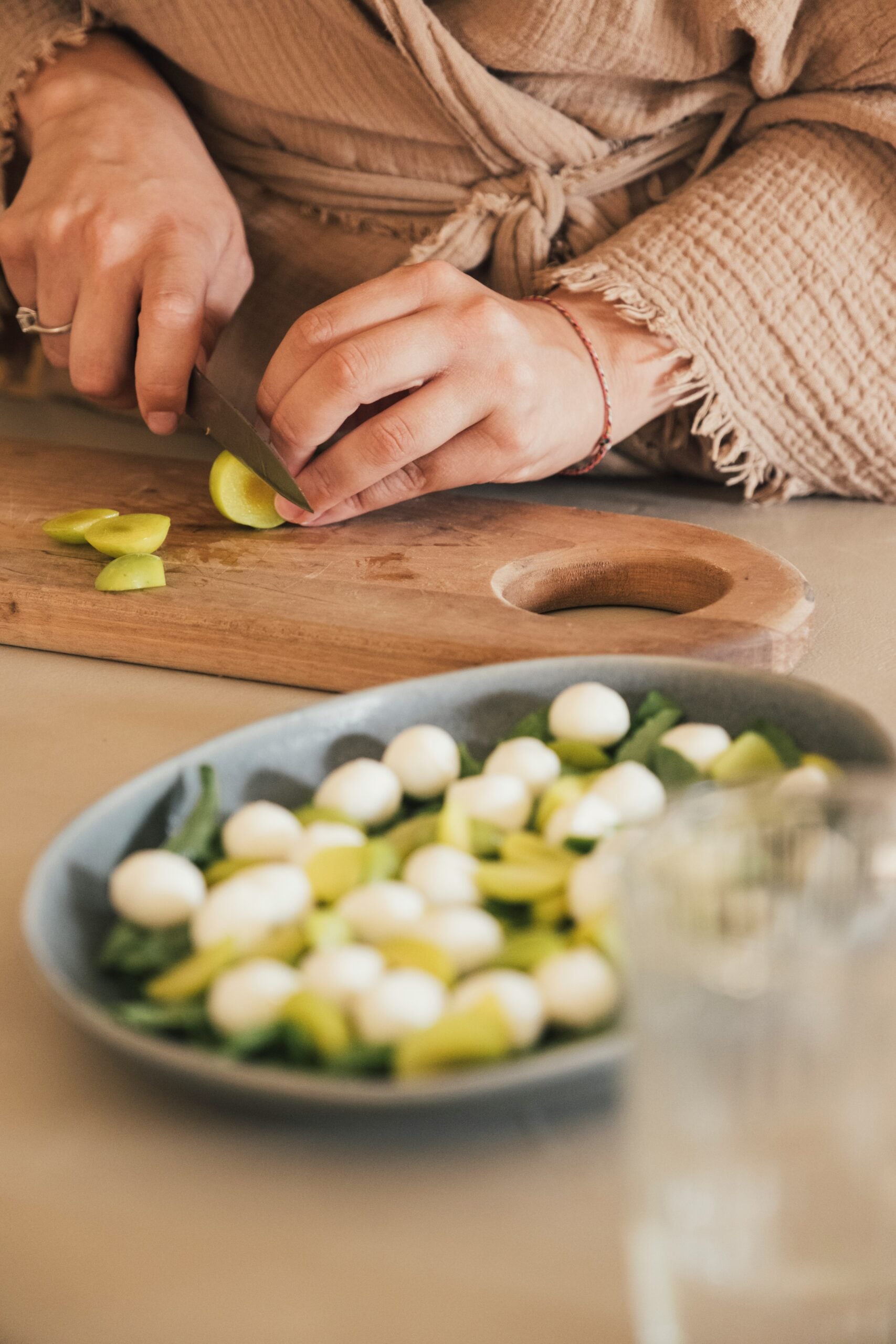 A person in a beige robe slices yellow tomatoes on a wooden cutting board. In the foreground, a plate holds mozzarella balls, spinach, and more yellow tomato slices.