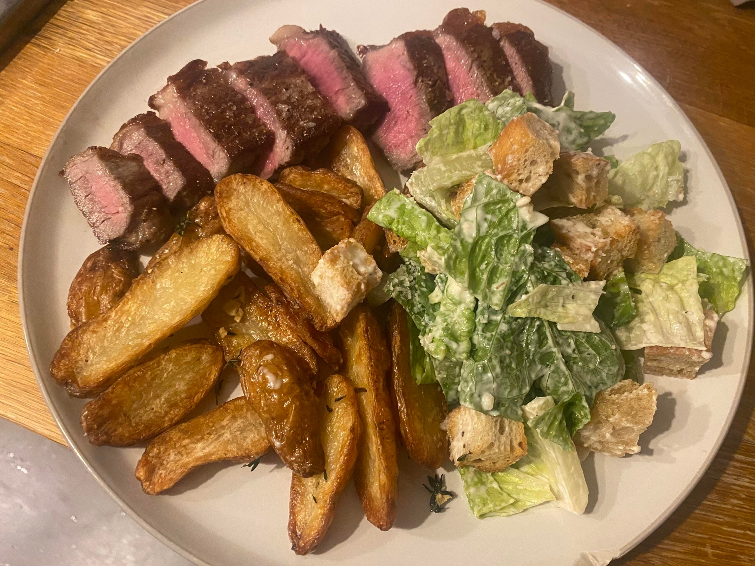 A plate with sliced medium-rare steak, crispy potato wedges, and Caesar salad with romaine, croutons, and creamy dressing.