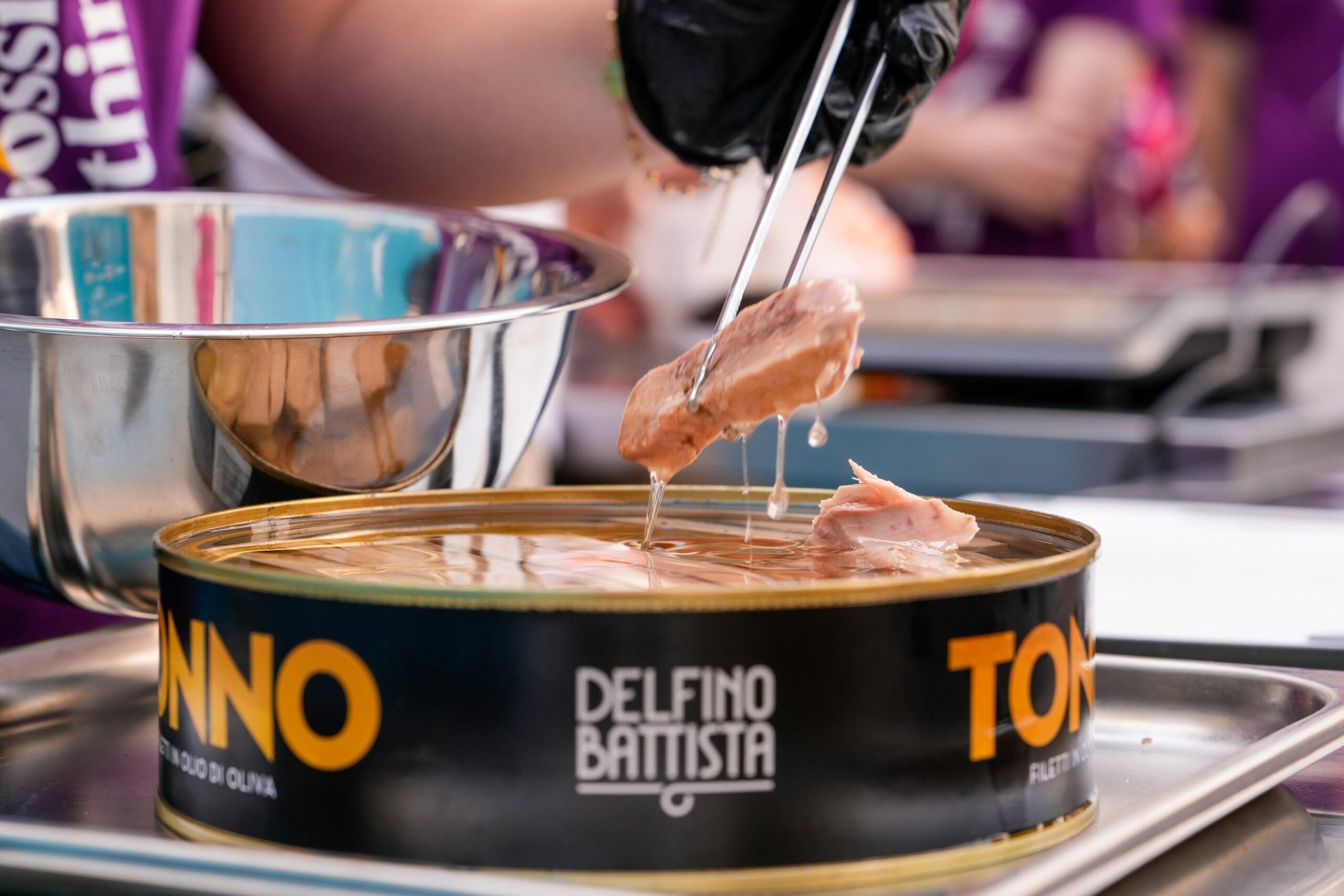 A person uses tongs to lift tuna pieces from a large open can labeled “Tonno Delfino Battista” during a food preparation event.