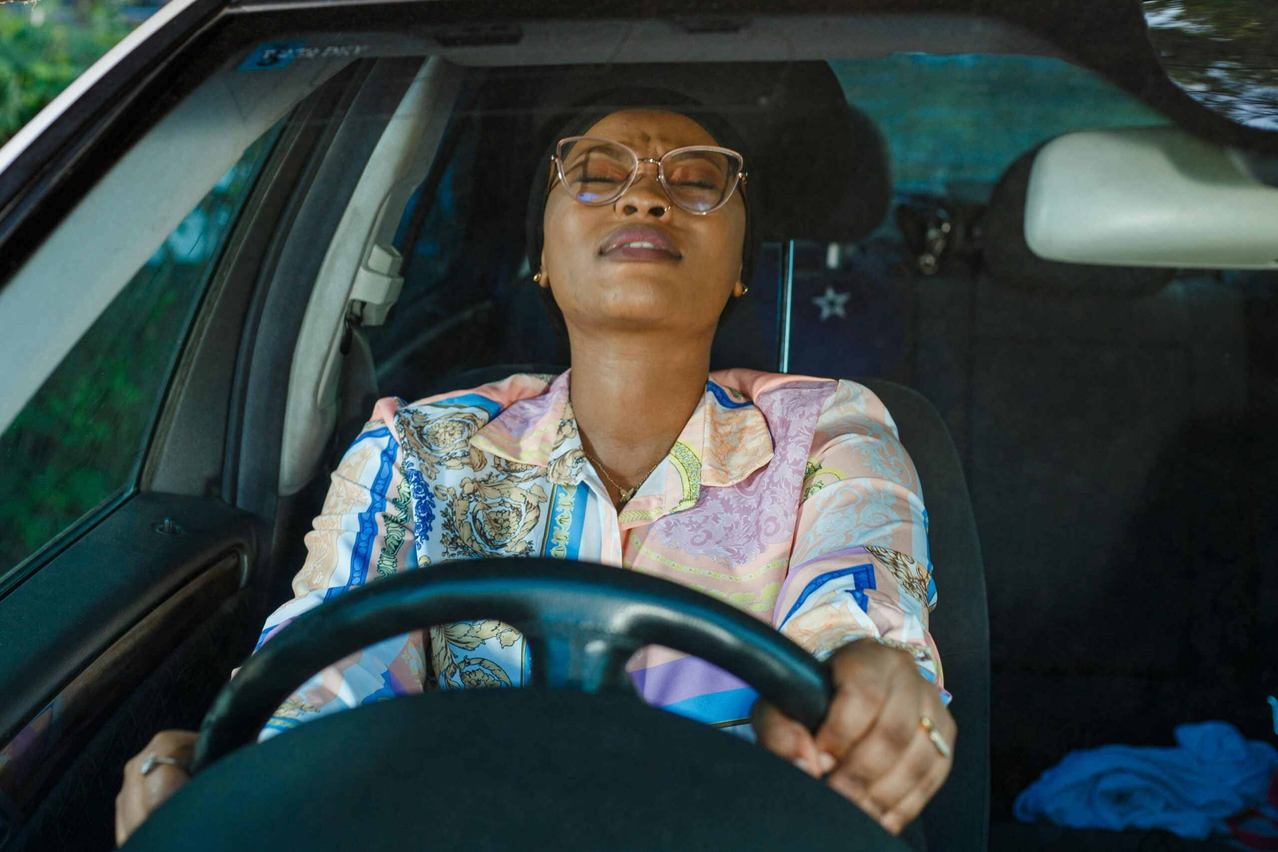 A woman sits in the driver’s seat of a car with her eyes closed and head tilted back, gripping the steering wheel tightly, appearing stressed or exhausted.