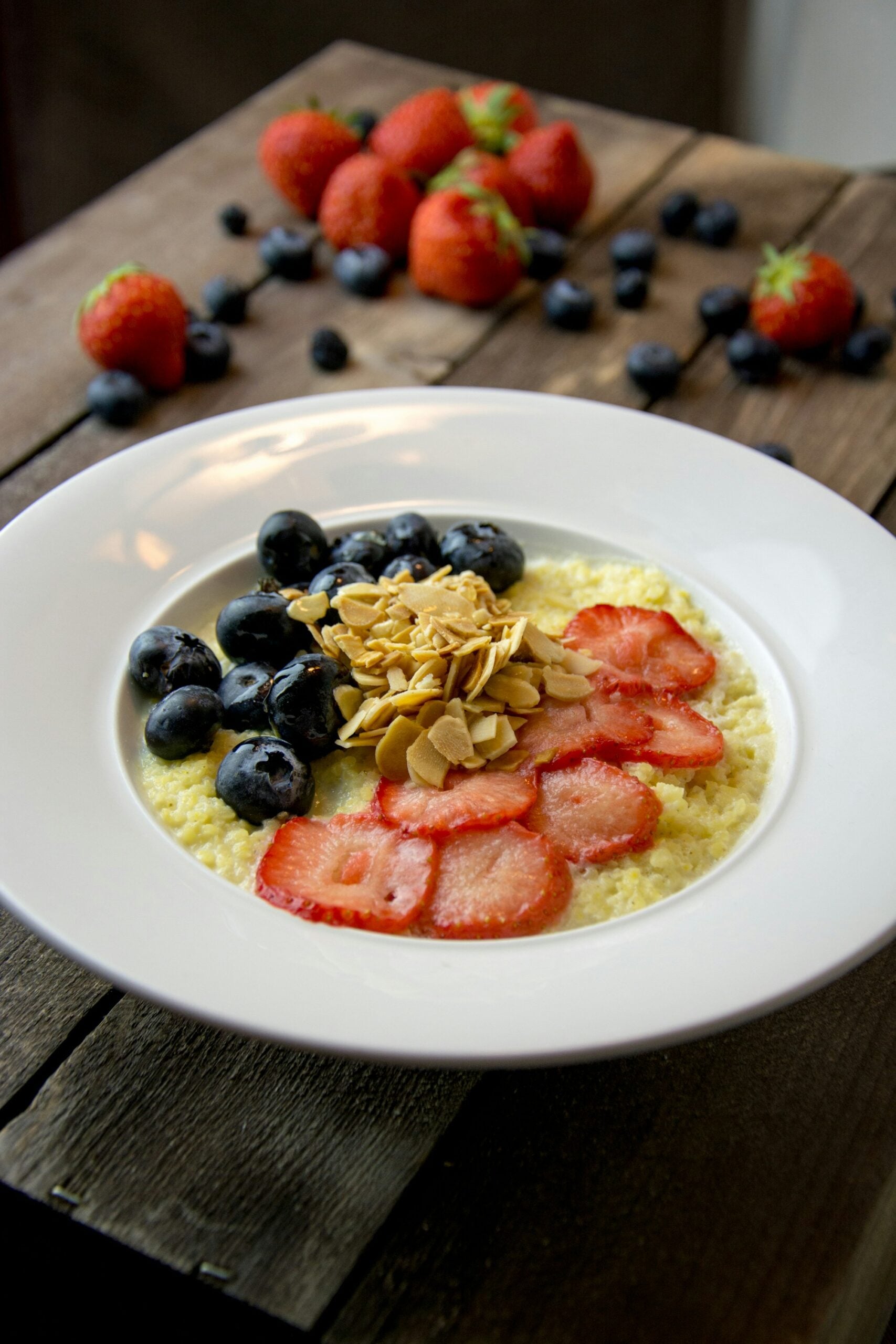 A white bowl contains oatmeal topped with sliced strawberries, blueberries, and almond slivers. Fresh strawberries and blueberries are scattered on a rustic wooden table in the background.