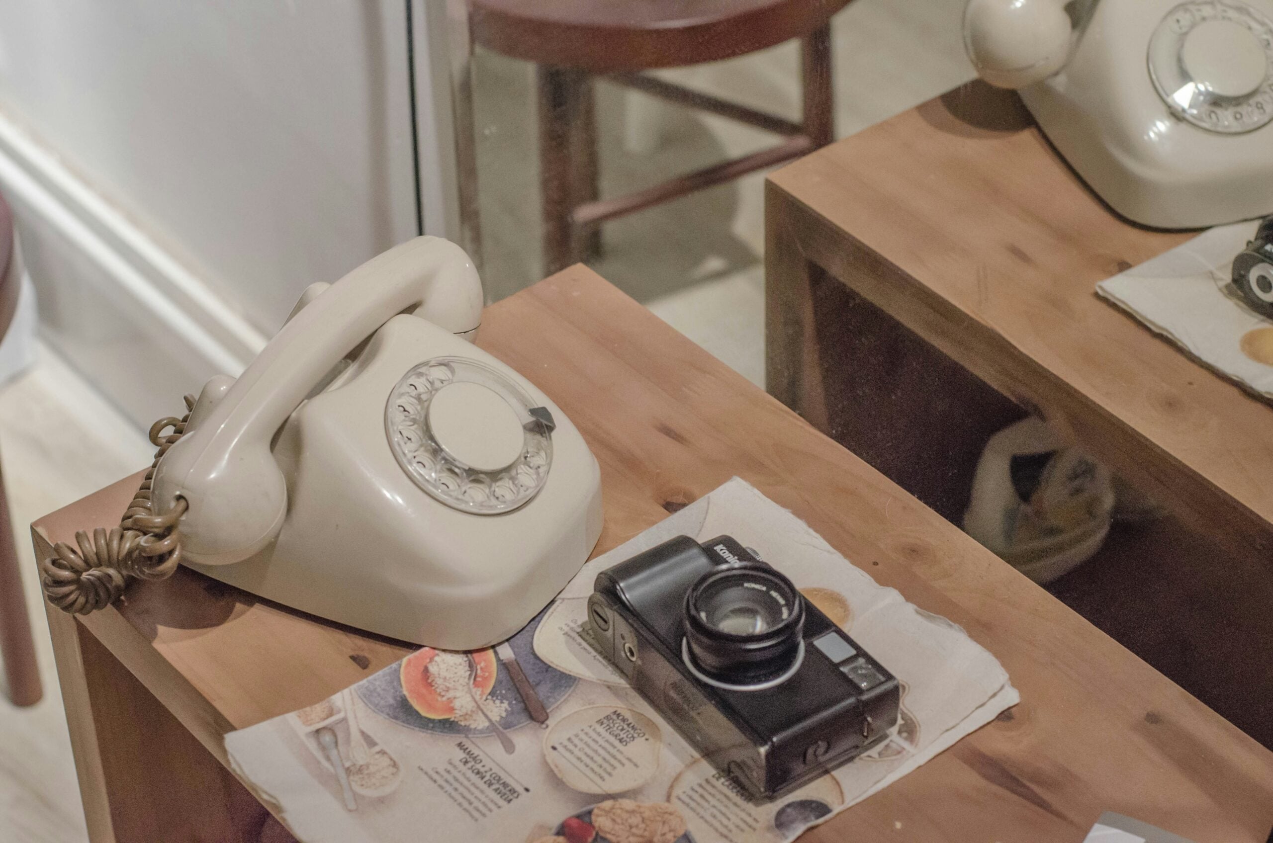 A beige rotary phone and a black vintage camera rest on a wooden table covered with a newspaper, reflected in a mirror beside a wooden chair.