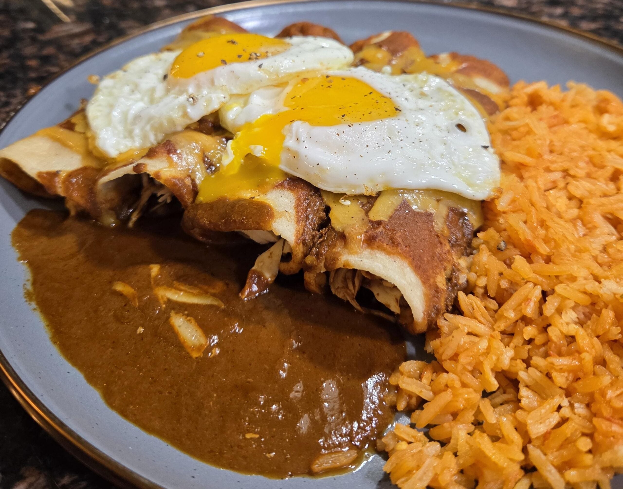 A plate of enchiladas topped with two sunny-side-up eggs and brown sauce, served with a side of Mexican rice.