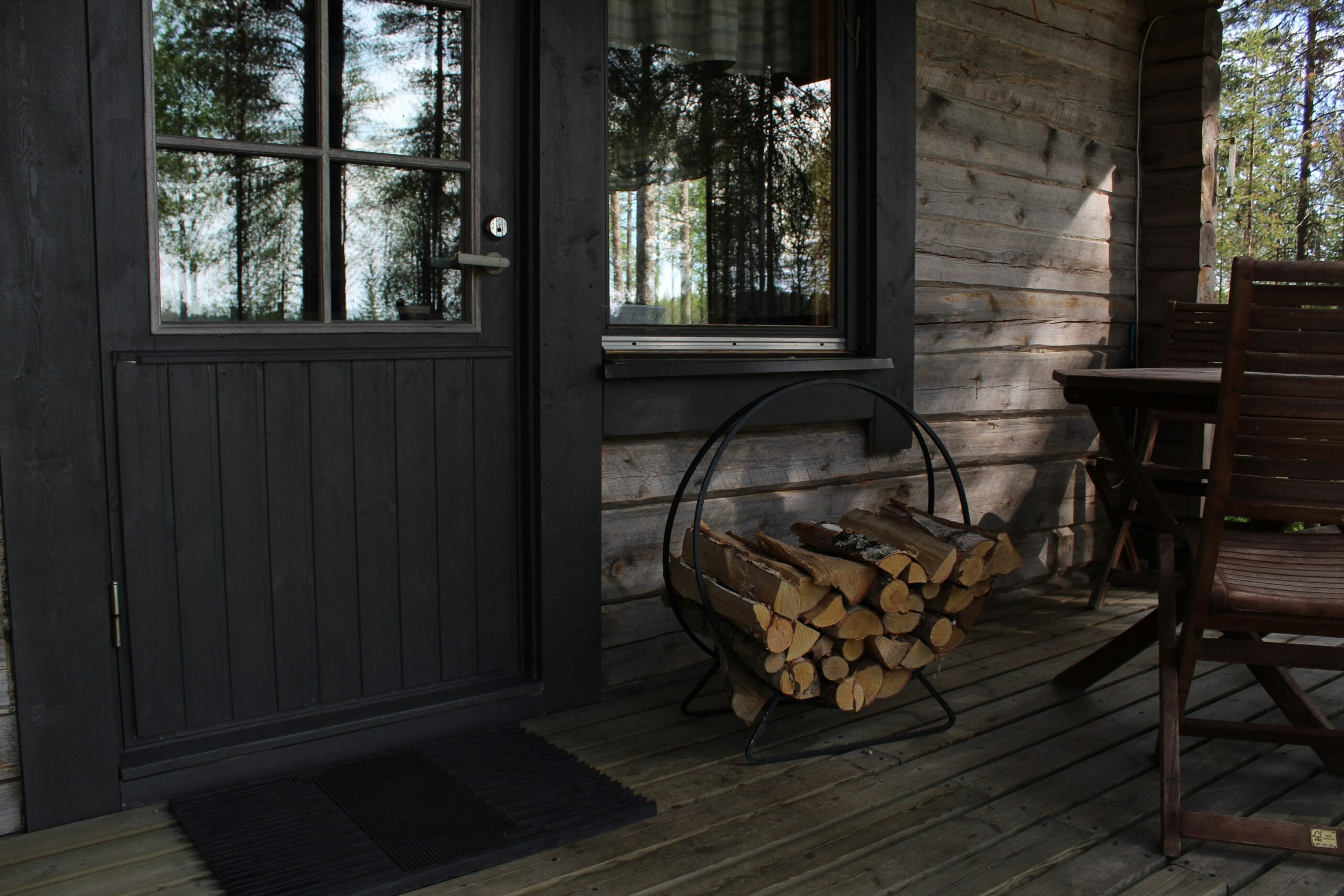 A stack of firewood in a metal holder sits on a wooden porch next to a dark door and window. Wooden chairs and a table are nearby; trees are reflected in the window and visible outside.