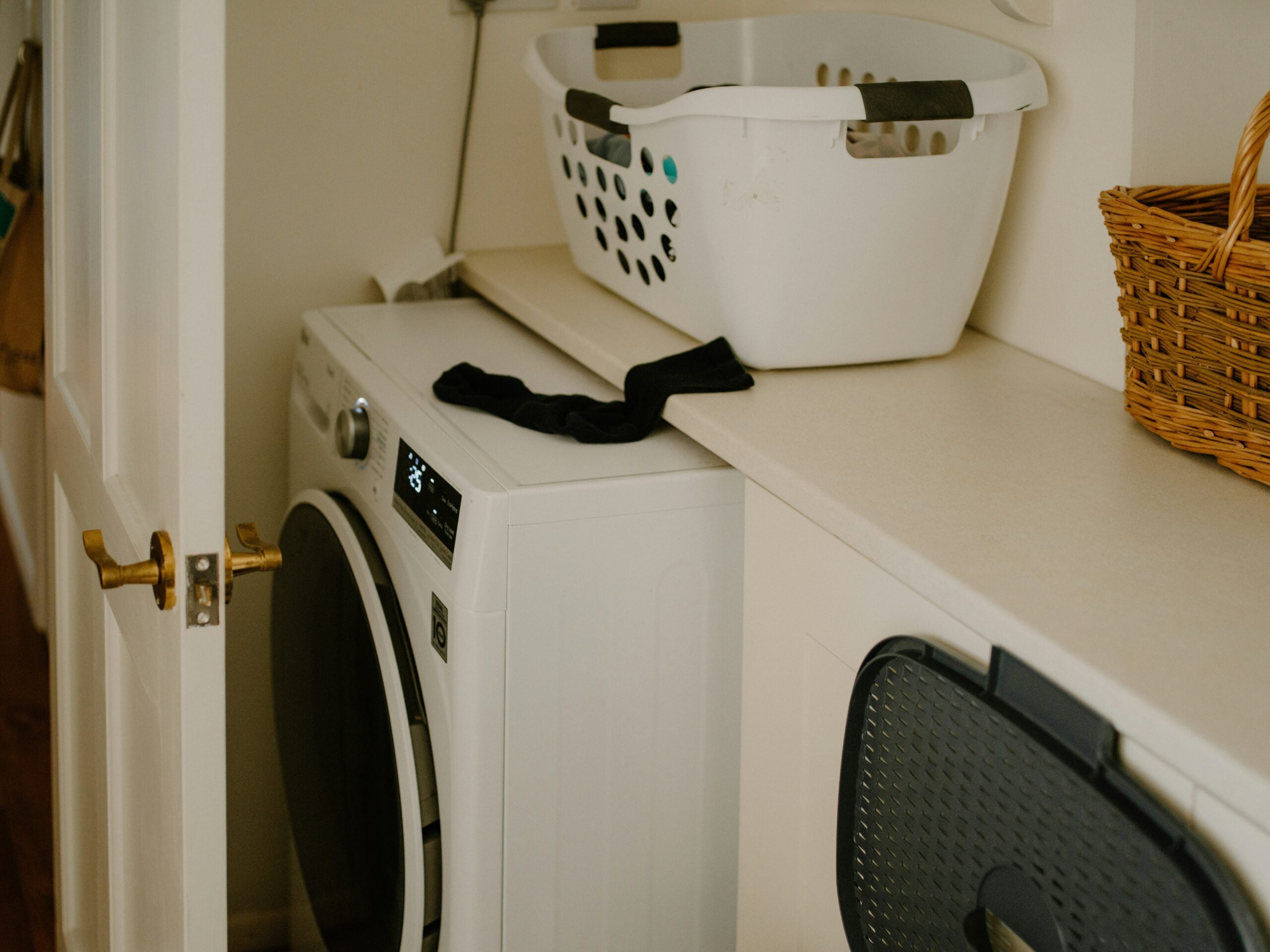 A laundry room with a front-load washing machine, a white laundry basket on the counter, a pair of black socks in front of it, a wicker basket nearby, and an open door partially blocking the view.
