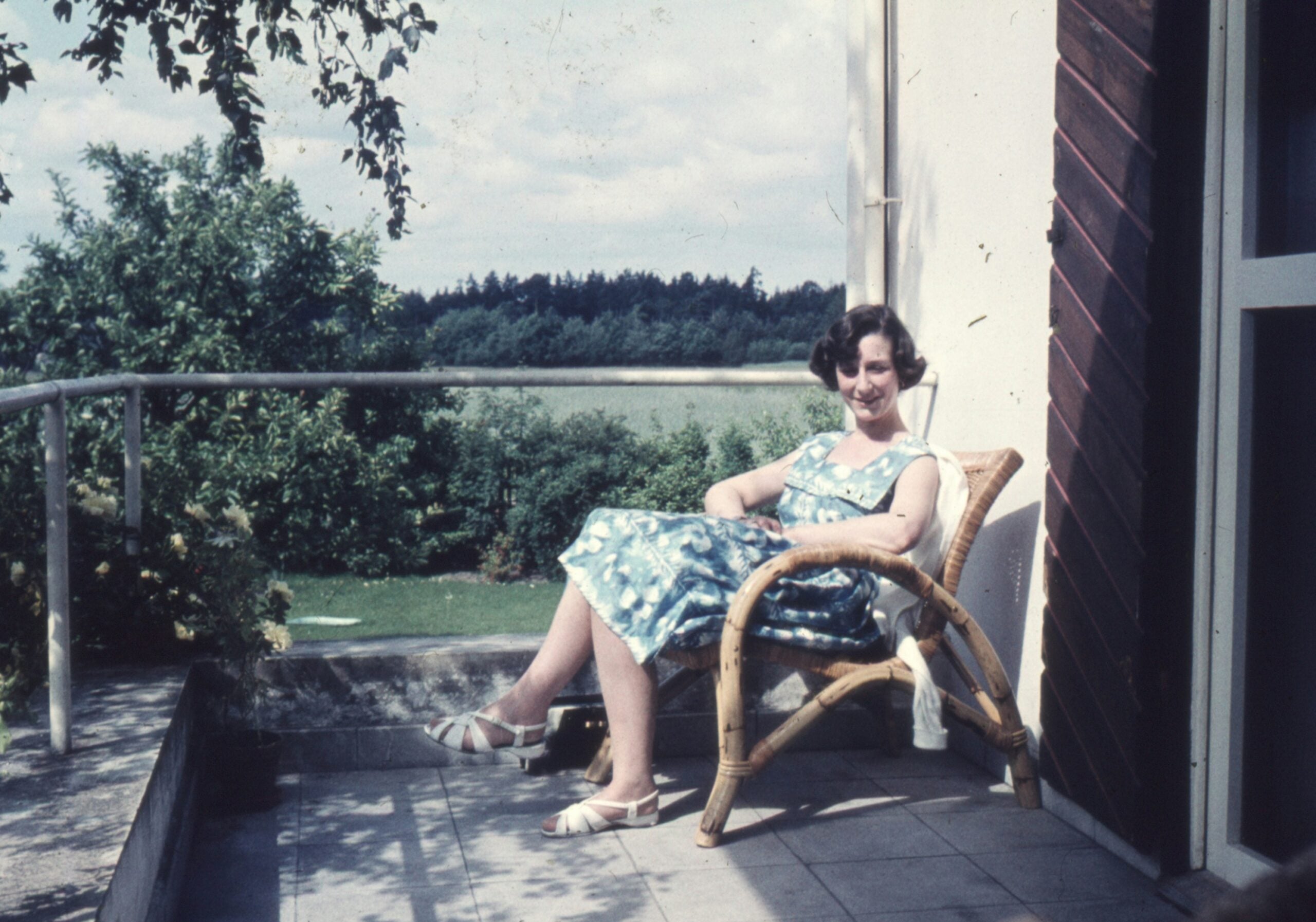 A woman in a blue dress sits on a wicker chair on a patio, smiling at the camera. Trees, grass, and a lake are visible in the background under a partly cloudy sky.