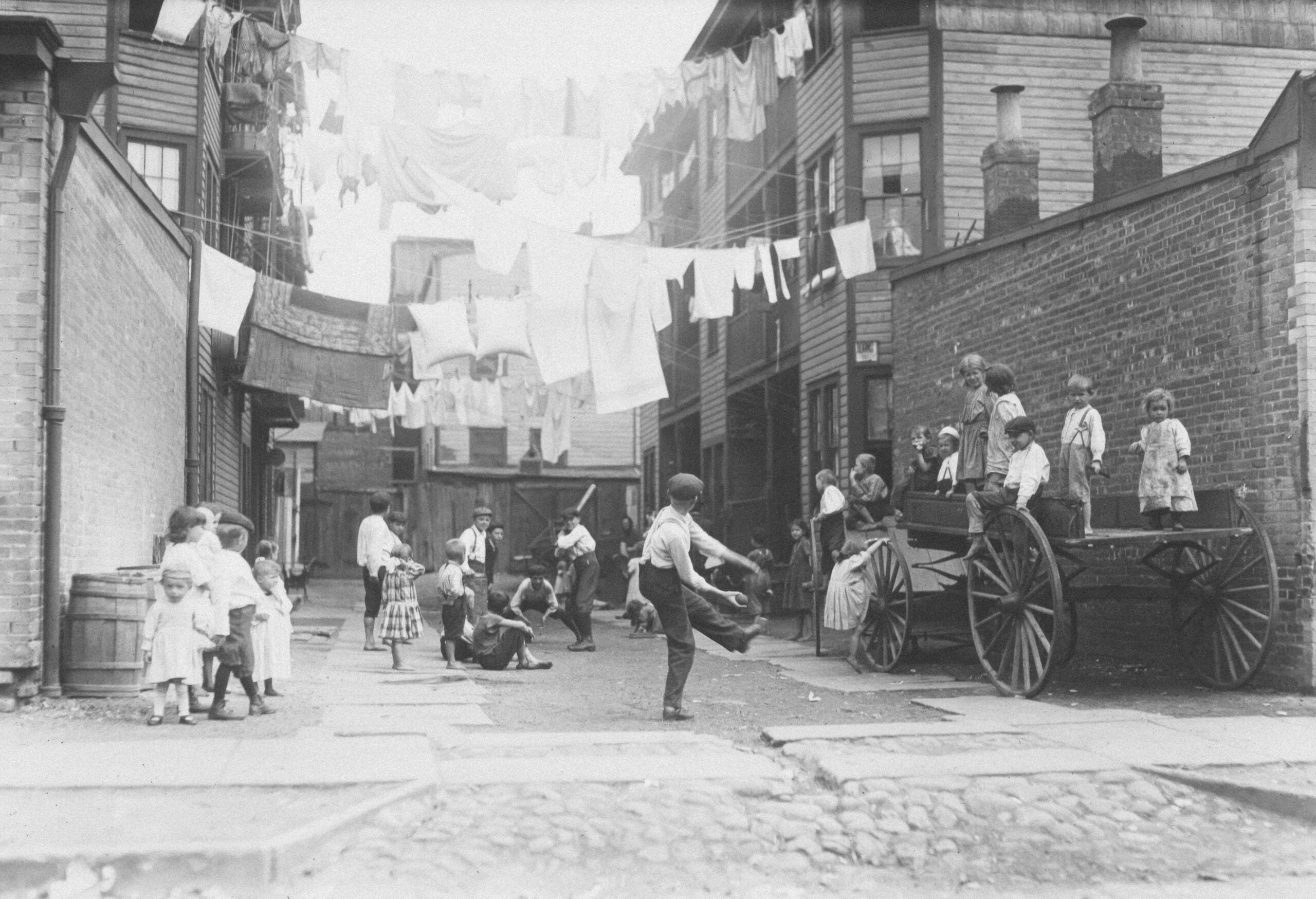 Black-and-white photo of children playing in a city alley, with some standing on a cart and others on the ground. Laundry hangs on lines above, and tall buildings line both sides of the alley.