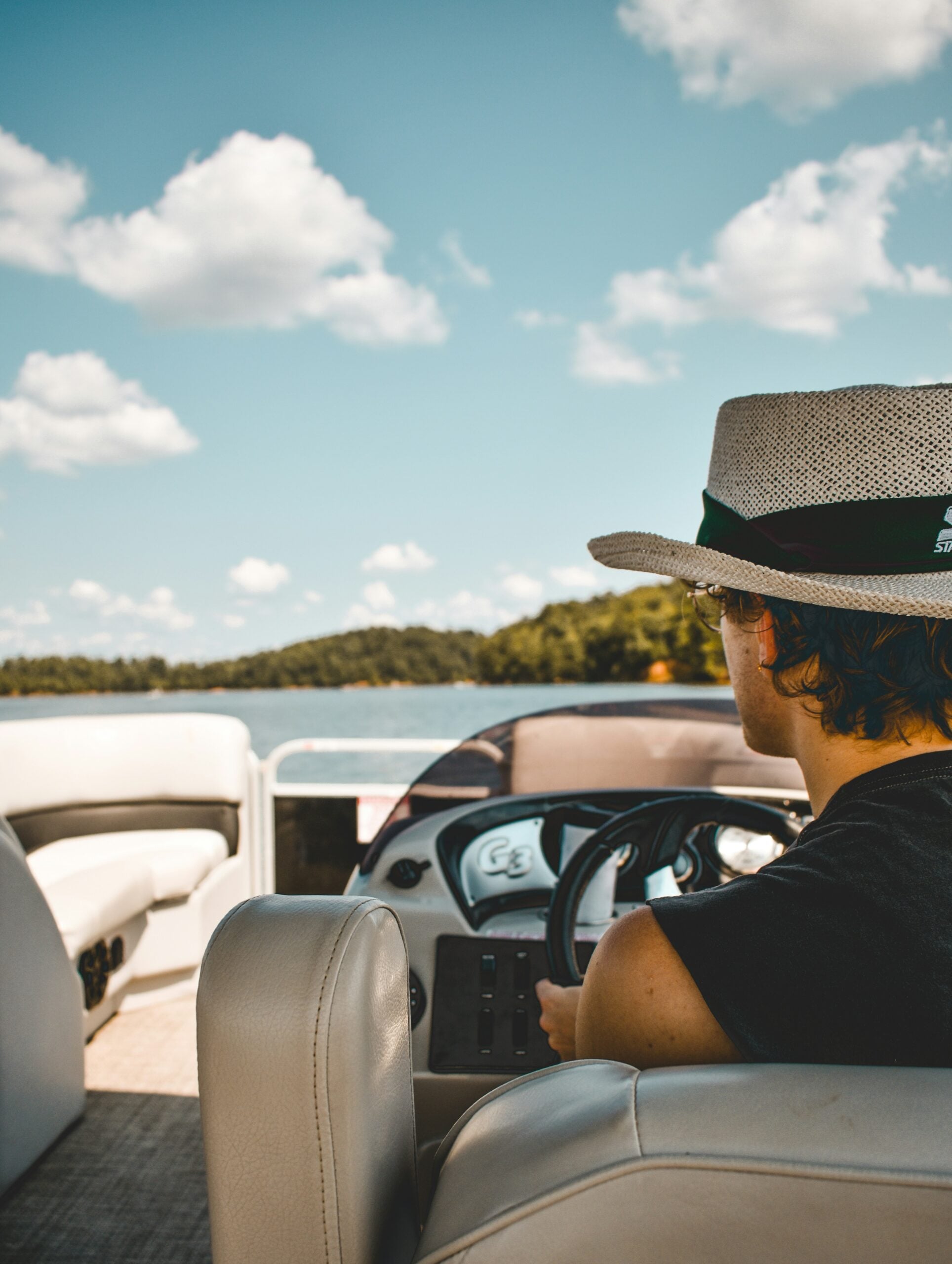 A person wearing a wide-brimmed hat steers a boat on a sunny day, with blue sky, scattered clouds, and trees visible in the background across the water.