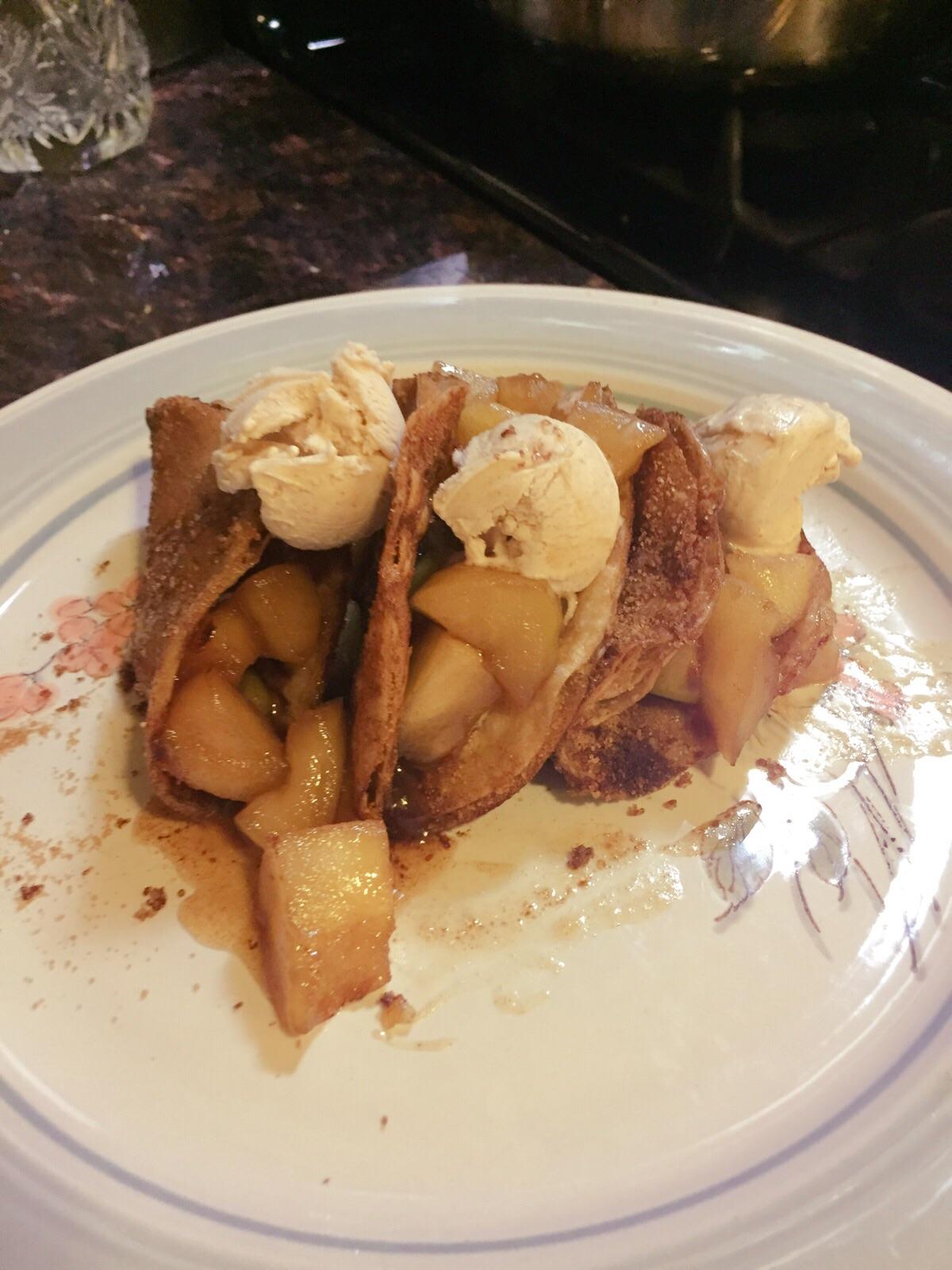Three cinnamon-sugar taco shells filled with cooked apples and topped with scoops of vanilla ice cream, served on a decorative plate.