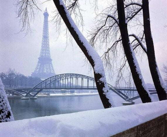 Snow-covered trees and a railing frame the Eiffel Tower and a bridge over the Seine River in Paris on a wintry day.