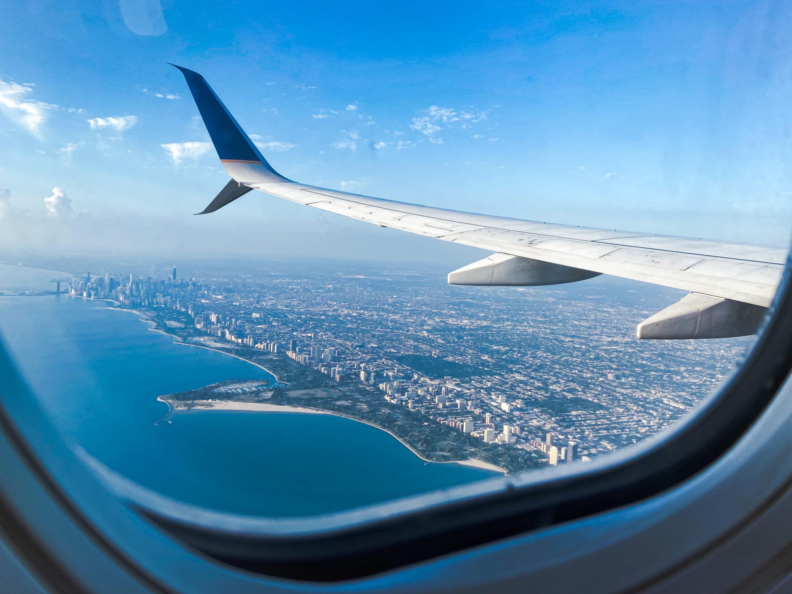 A view from airplane windows reveals the wing, a city skyline, and the coastline below under a clear blue sky.