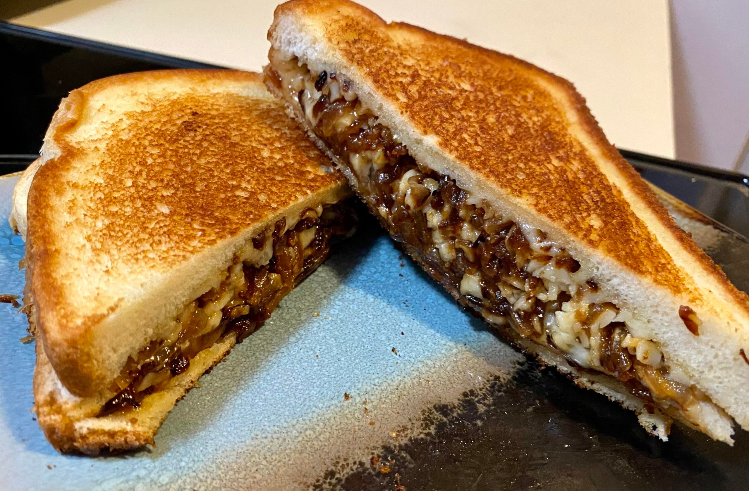 Two halves of a toasted Grilled Cheese Sandwich filled with melted cheese and caramelized onions are displayed on a blue plate. The golden brown bread looks crispy, and the filling is visible in the cross-section.