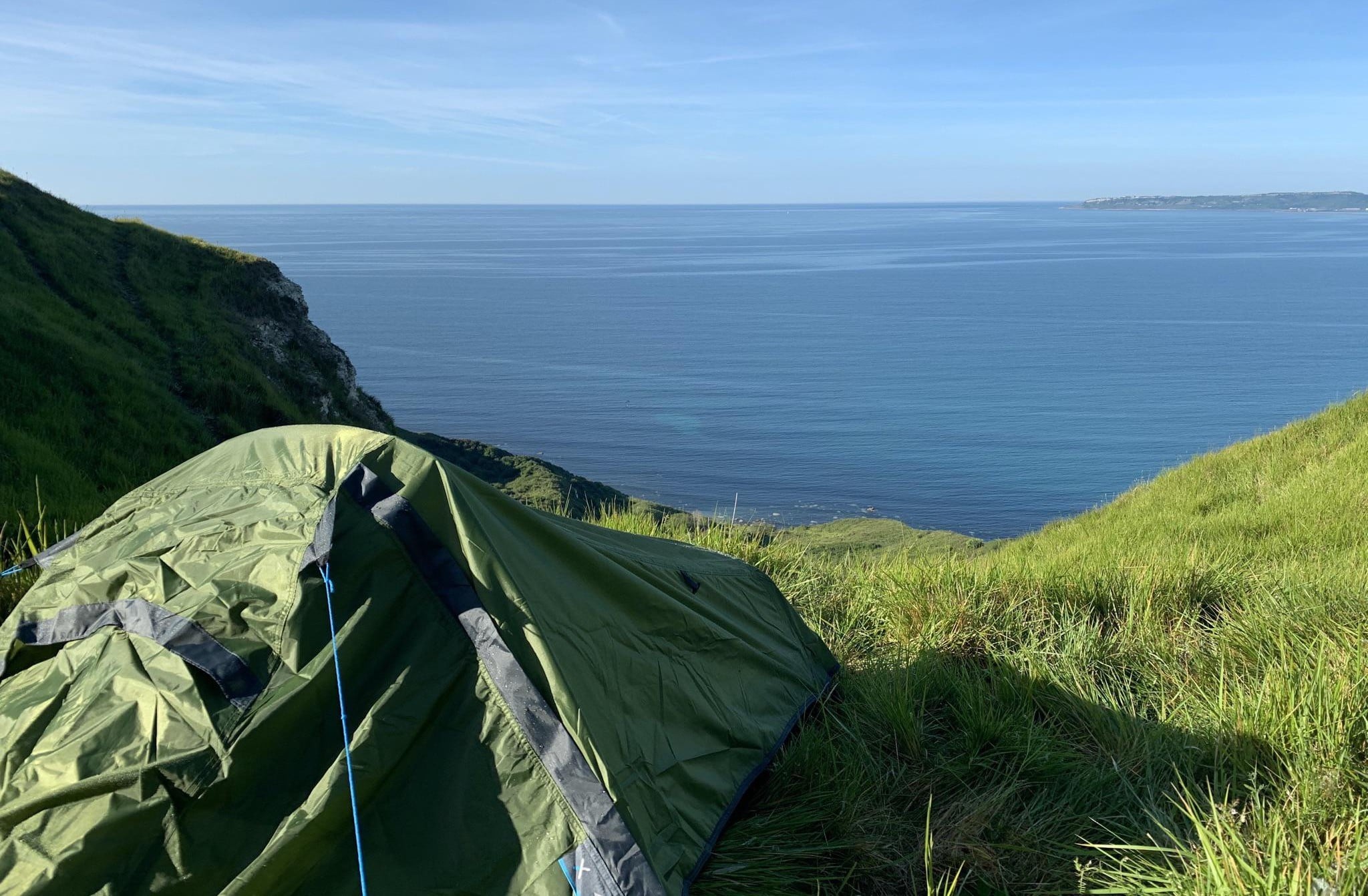 A green tent is pitched on a grassy slope overlooking a calm, blue sea under a clear sky, with distant coastline visible on the horizon.
