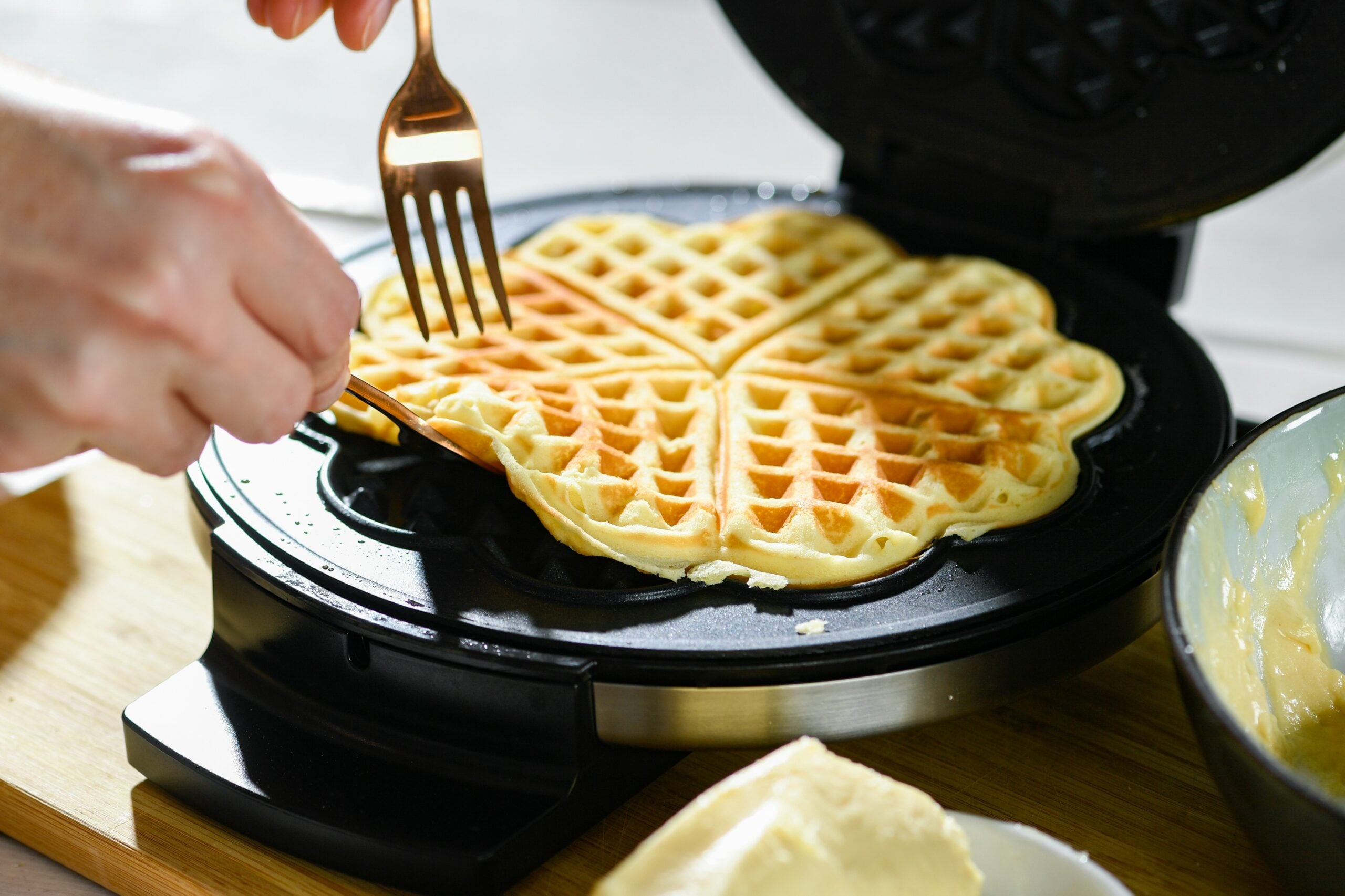 A hand uses a fork to lift a freshly cooked, golden, heart-shaped waffle from a waffle maker, with batter and butter visible nearby on a wooden surface.