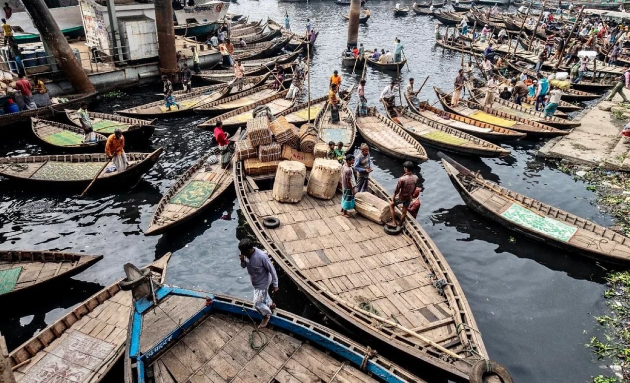 Numerous wooden boats crowded together on a busy river, with people loading goods and moving between vessels; the vibrant, lively atmosphere with baskets and market activity is reminiscent of the bustling waterways found in some of the Biggest Cities In The World.