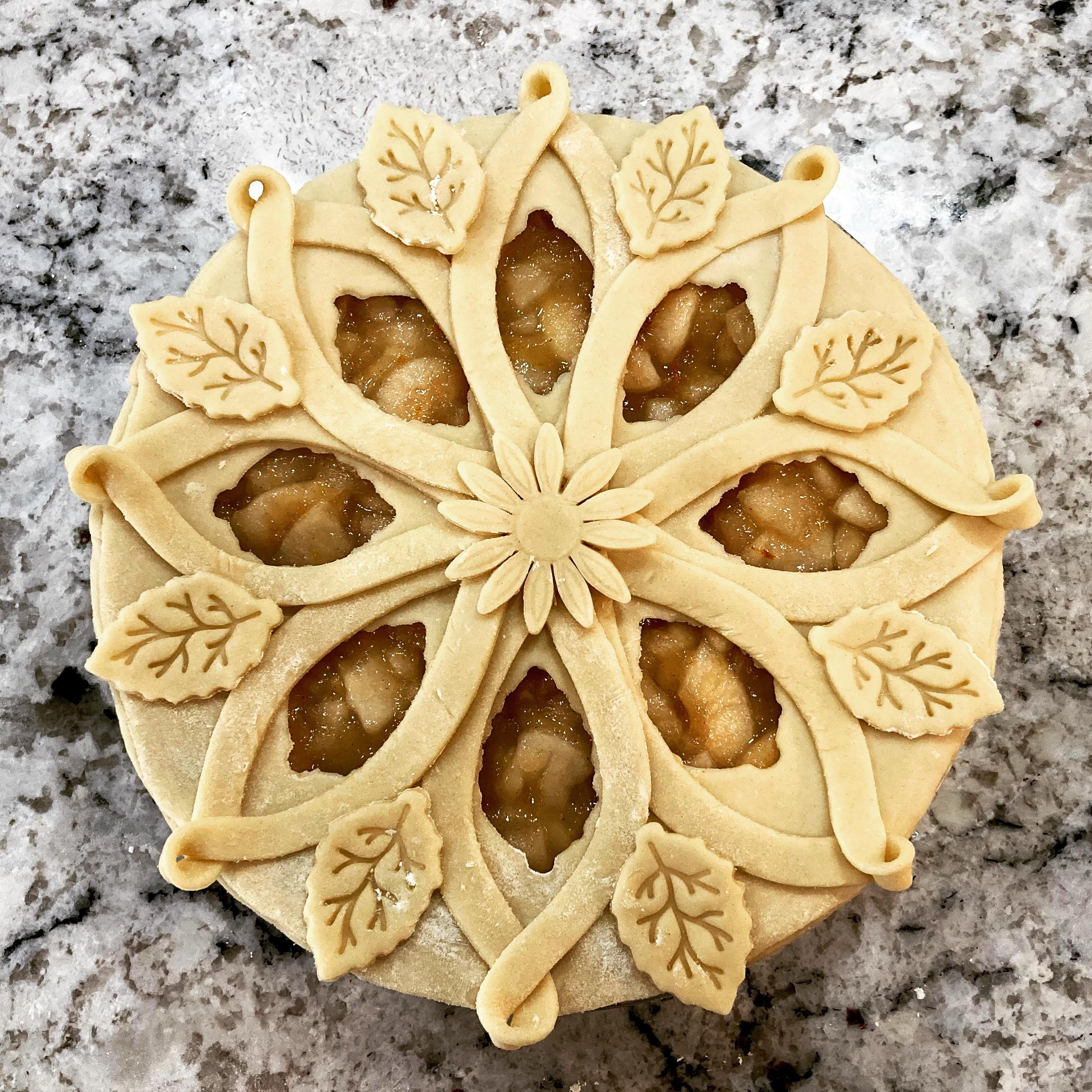 Unbaked pie with an intricate, decorative top crust featuring leaf and floral patterns, revealing apple filling through petal-shaped cutouts. The pie rests on a marbled countertop.