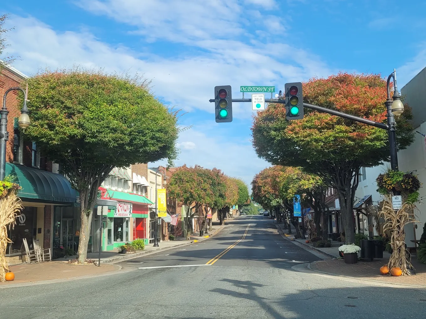 A small town street lined with trees and shops, decorated with cornstalks and pumpkins. The traffic light is green, and the sky is mostly clear with a few clouds. Affordable mountain towns for retirement.