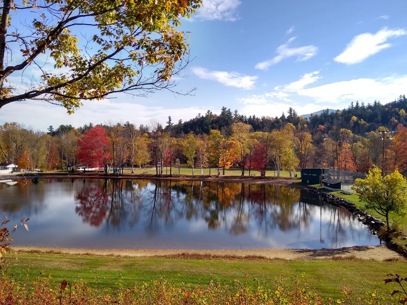 A calm pond reflects colorful autumn trees under a blue sky, with a sandy shore and green grass in the foreground. A small building is visible near the water.