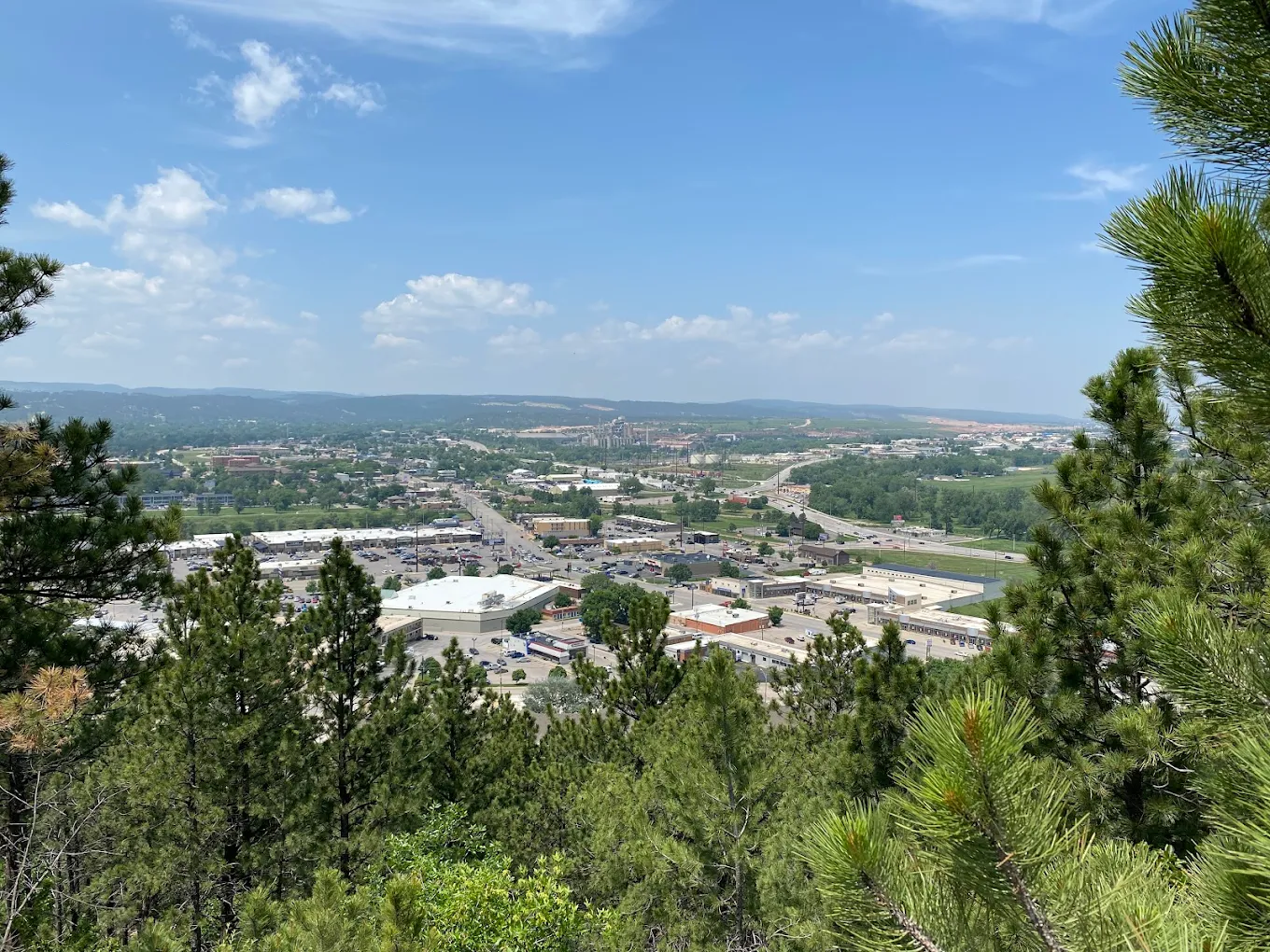 A cityscape seen from a hillside surrounded by pine trees, with buildings, roads, and green areas under a blue sky with scattered clouds.