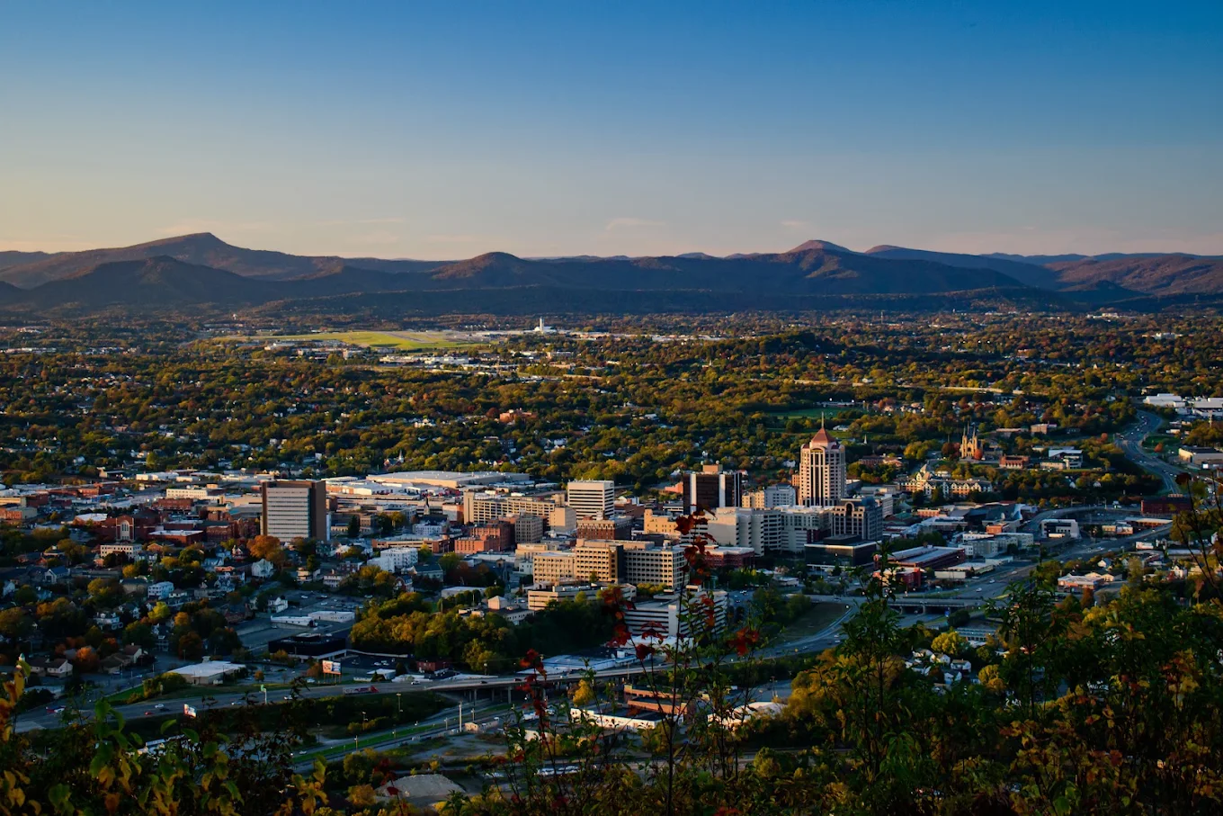 Cityscape at sunset with downtown buildings, roads, and green trees, set against rolling mountains and a clear blue sky in the background.