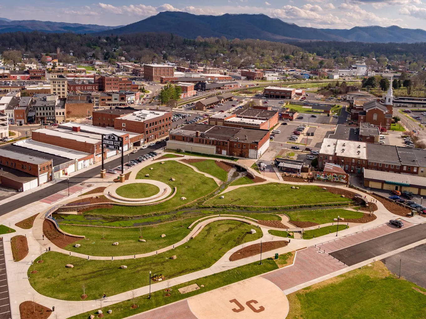 Aerial view of a city park with winding paths, circular plazas, and green lawns, surrounded by buildings and roads, with mountains in the background under a partly cloudy sky. “JC” is marked on the pavement in the foreground.