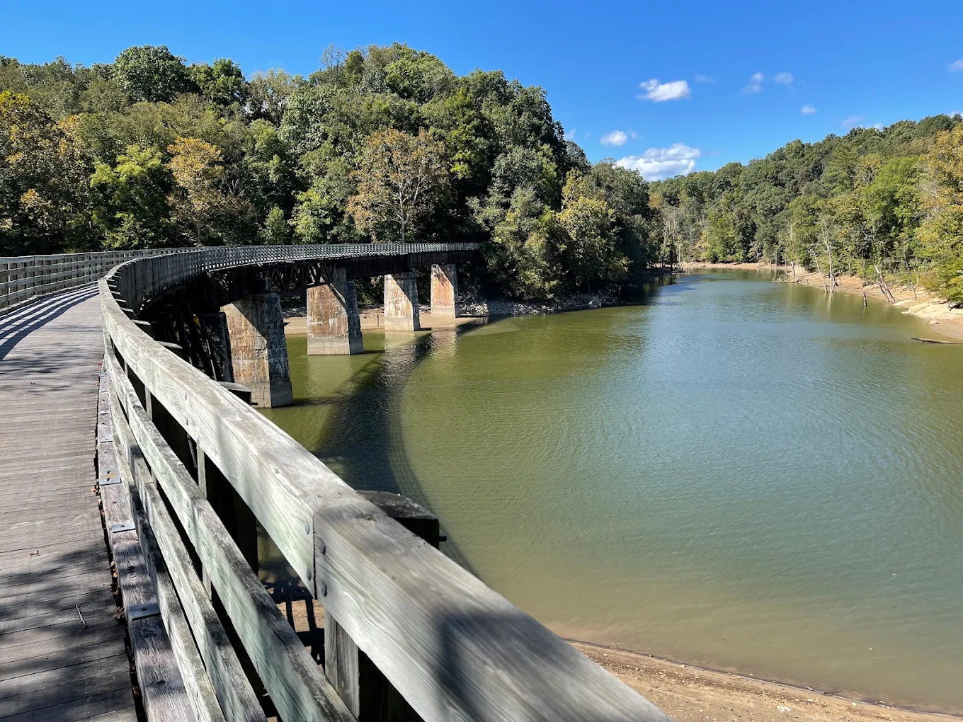 Curved wooden bridge with railings spans over a calm river, surrounded by dense green trees under a clear blue sky, on a sunny day.