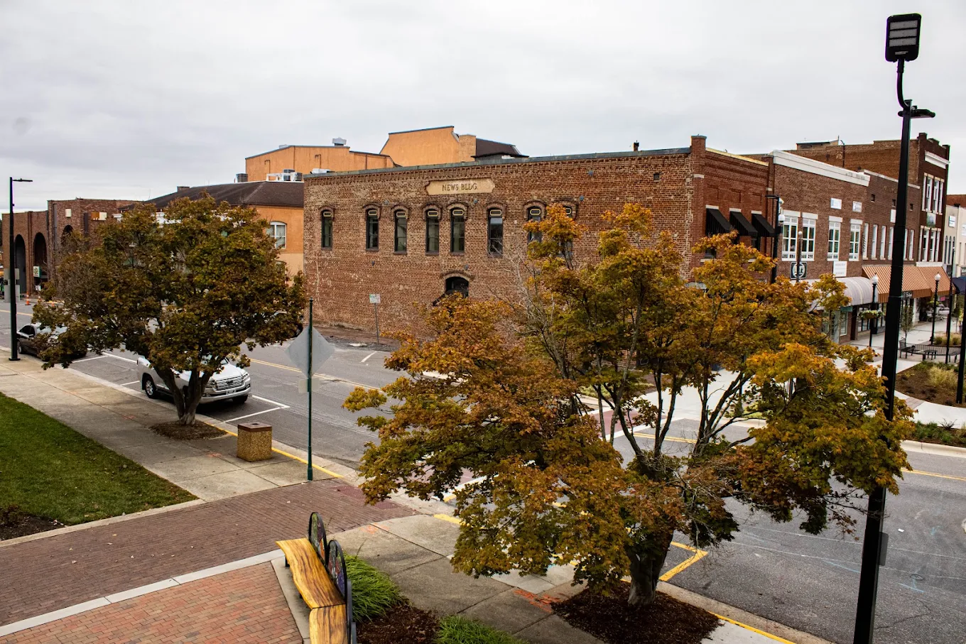 A small town street with brick buildings, trees with autumn leaves, a white pickup truck parked on the side, and cloudy skies overhead.
