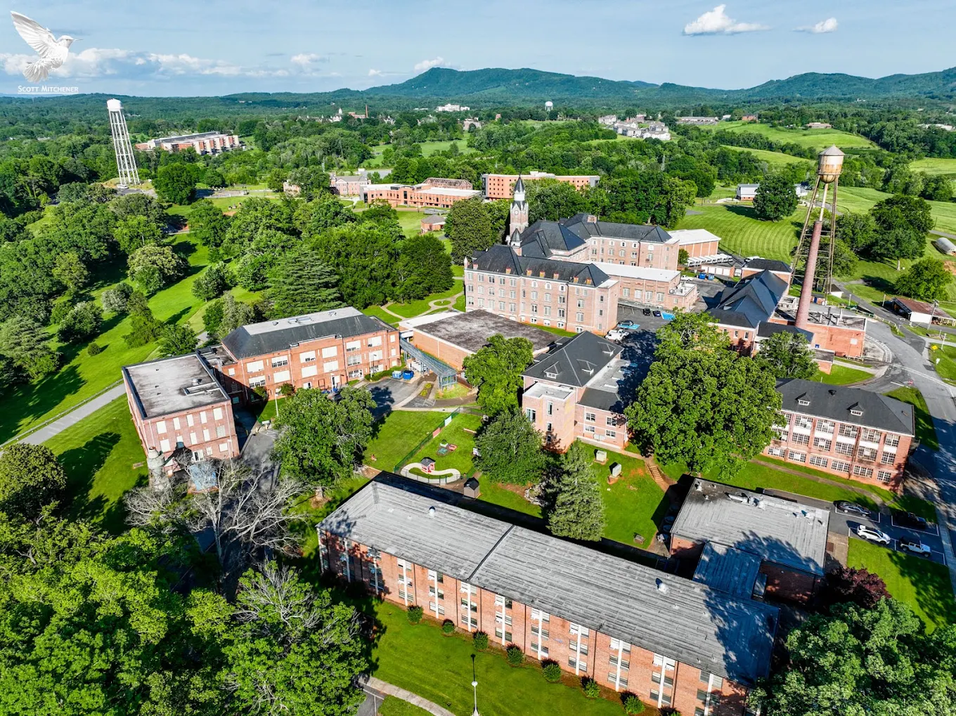 Aerial view of a college campus with multiple brick buildings, green trees, water towers, and distant hills under a blue sky with scattered clouds.