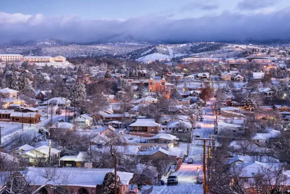 A snowy town at dusk with houses, trees, and a church, all covered in snow. The streets are lined with glowing lights, and mountains are visible in the background under a cloudy sky.