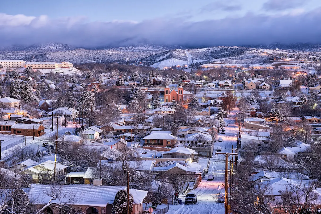 A snowy town at dusk with houses, trees, and a church, all covered in snow. The streets are lined with glowing lights, and mountains are visible in the background under a cloudy sky.